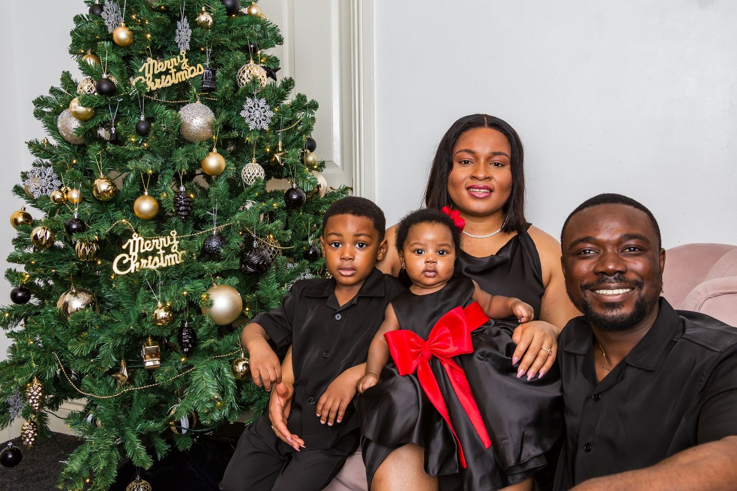 A joyful family of four poses by a decorated Christmas tree, wearing matching black and red outfits.