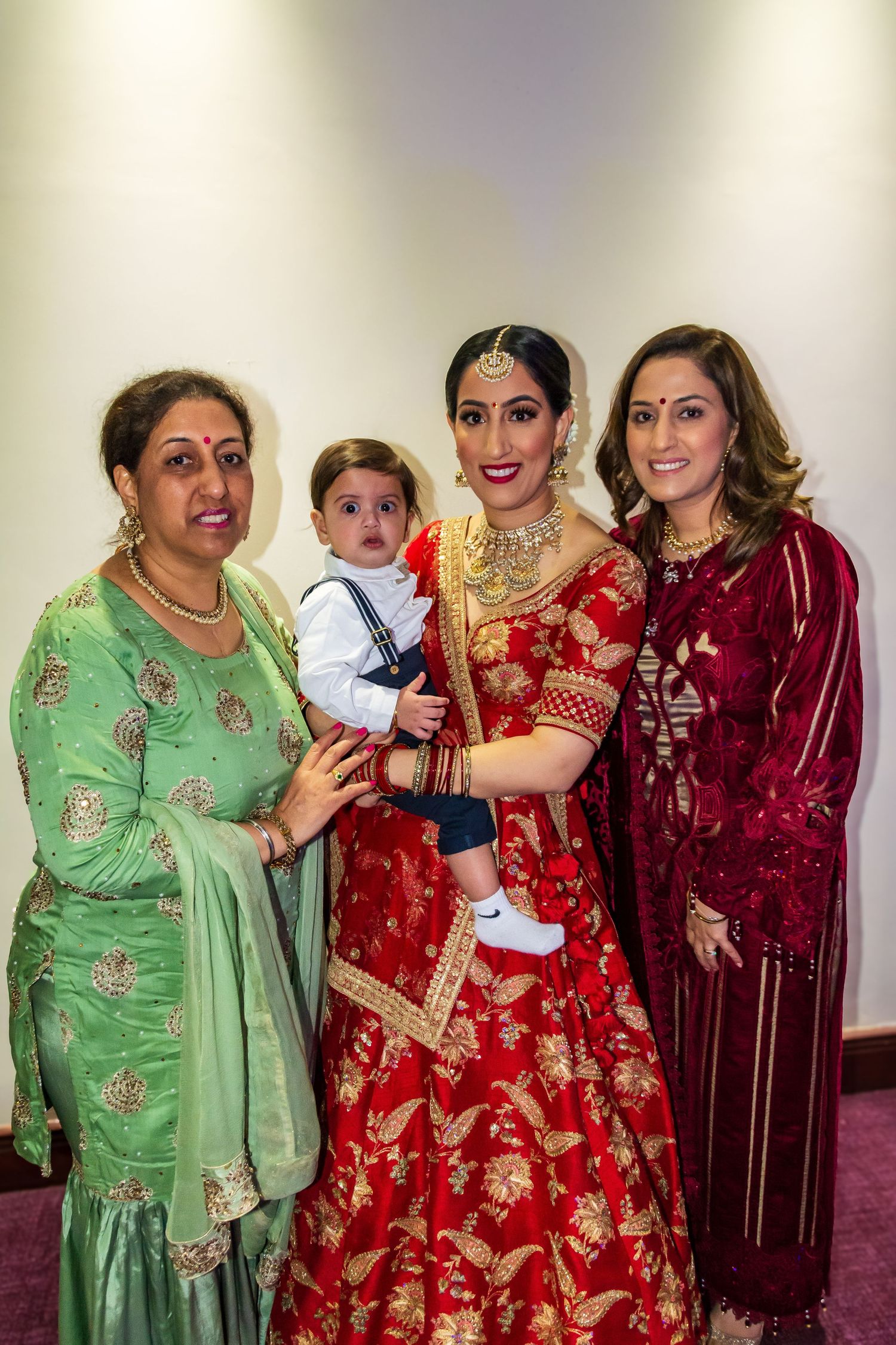 Bride in red lehenga holding baby with two women in traditional Indian attire at wedding.