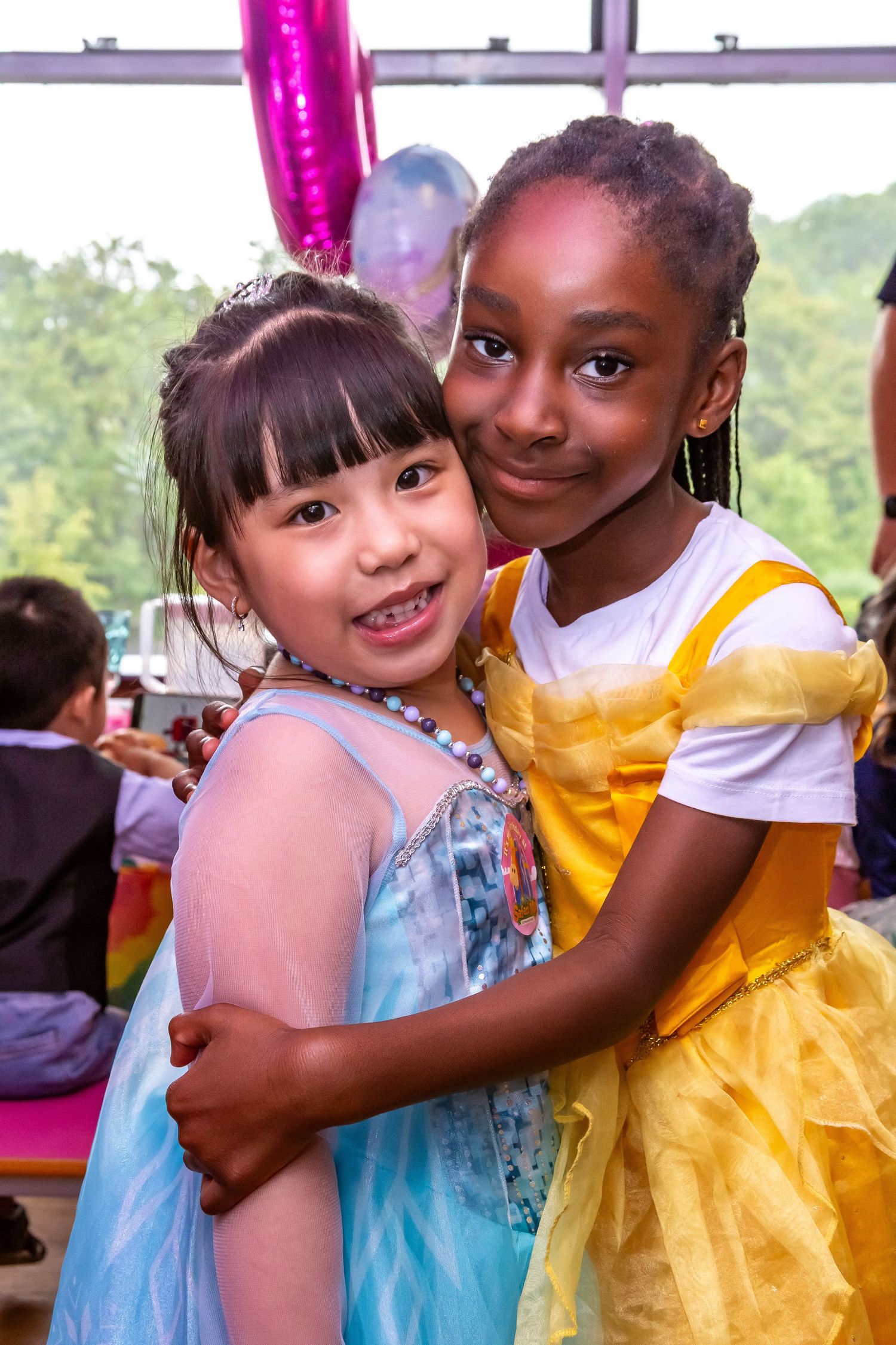 Two young girls in princess costumes hugging and smiling at a birthday party.
