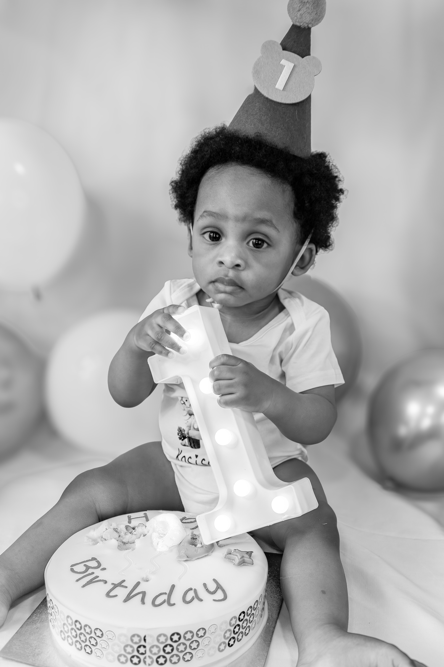 Black and white photo of baby boy in birthday hat with cake and number one sign at first birthday party.