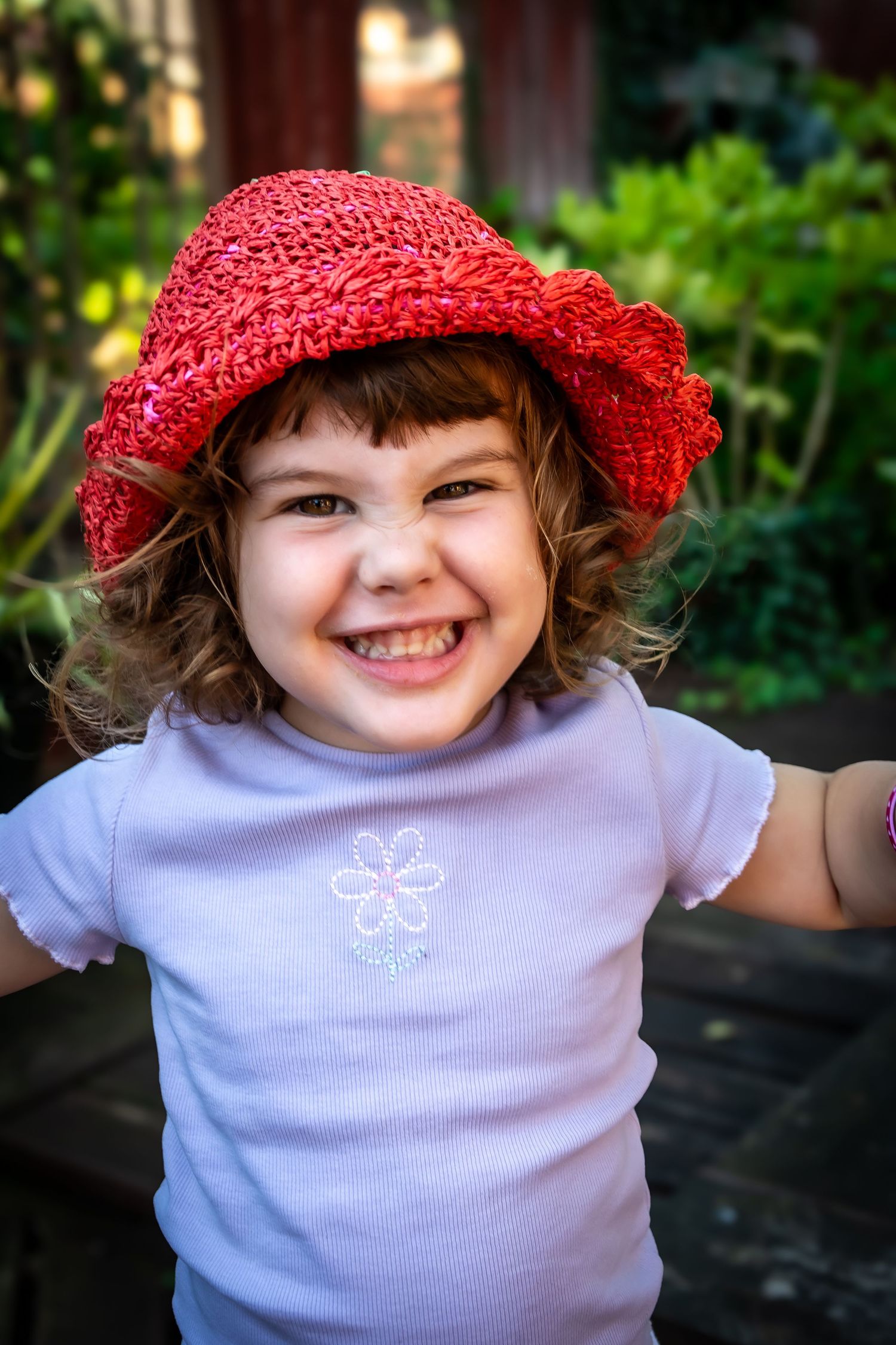 A cheerful young girl wearing a red straw hat and white floral t-shirt smiles brightly in a garden.