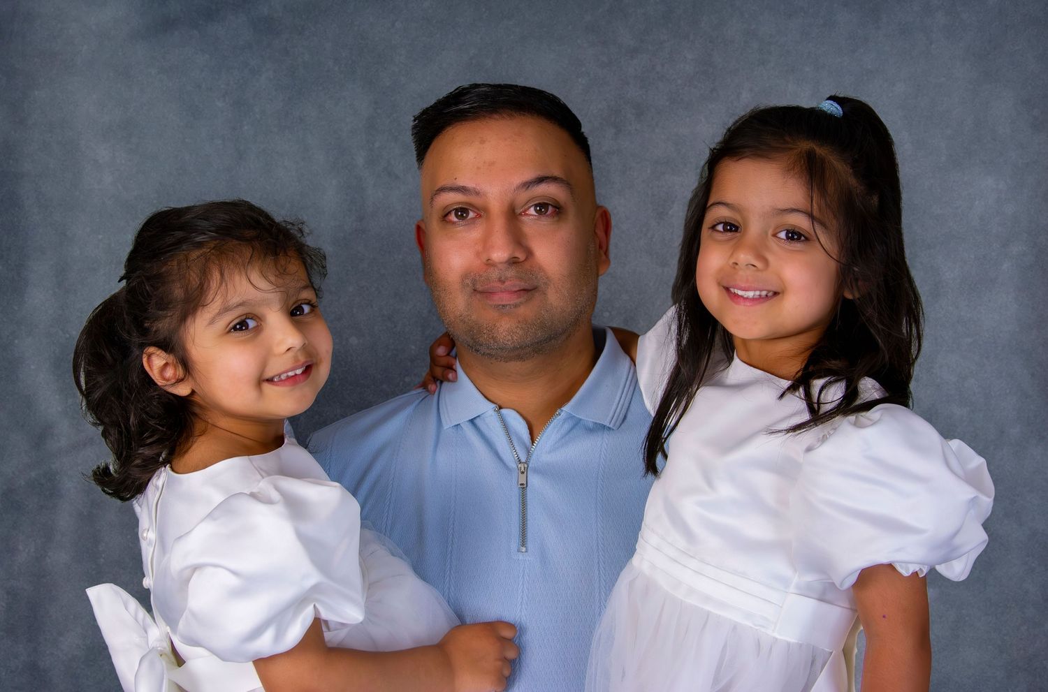 Father in blue shirt posing with two young daughters in white dresses for studio portrait.