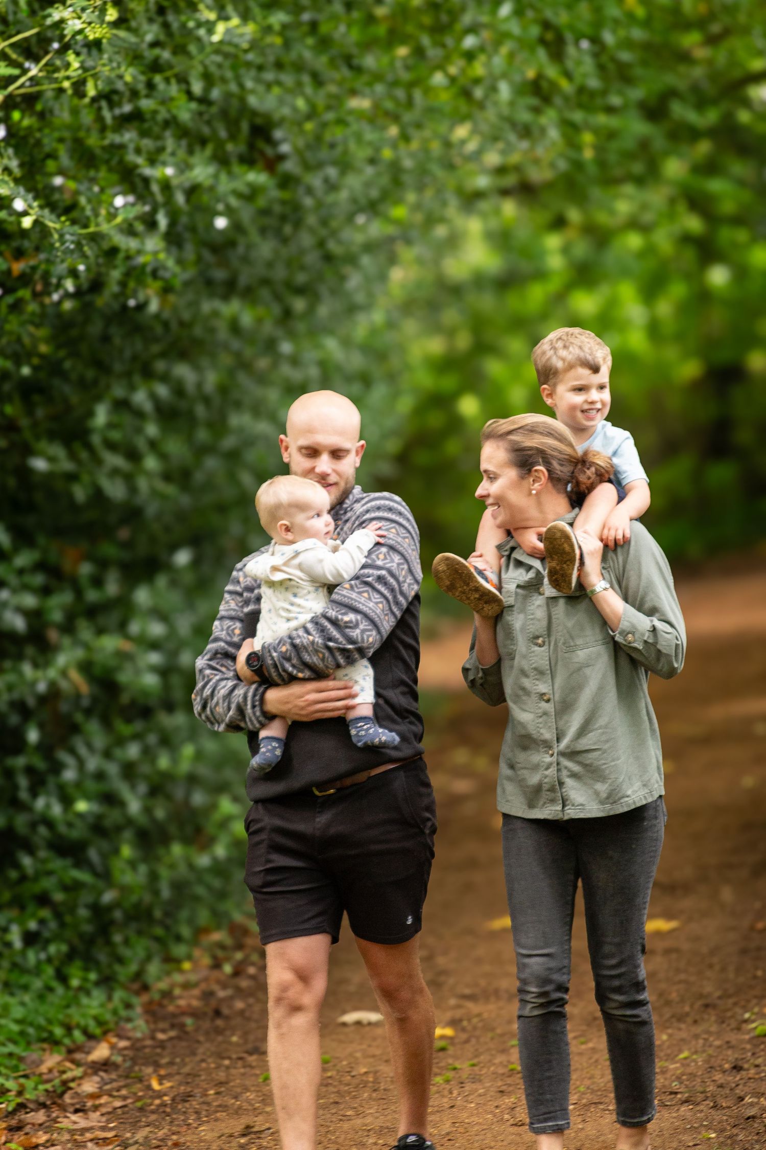 A happy family walking on a forest path, parents carrying two young children surrounded by lush green trees.