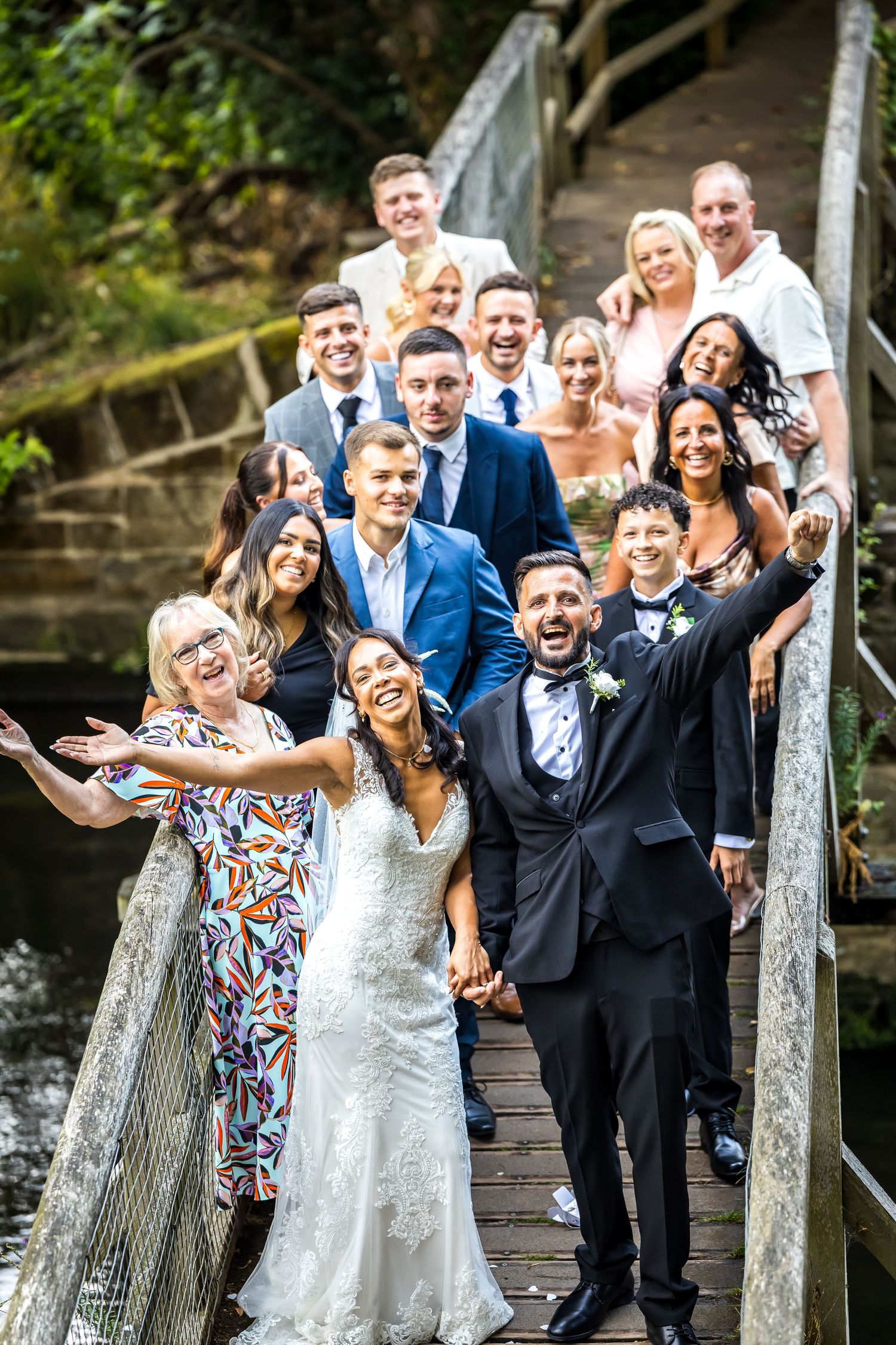 A joyful wedding party poses on a wooden bridge, with the bride and groom celebrating in the foreground.