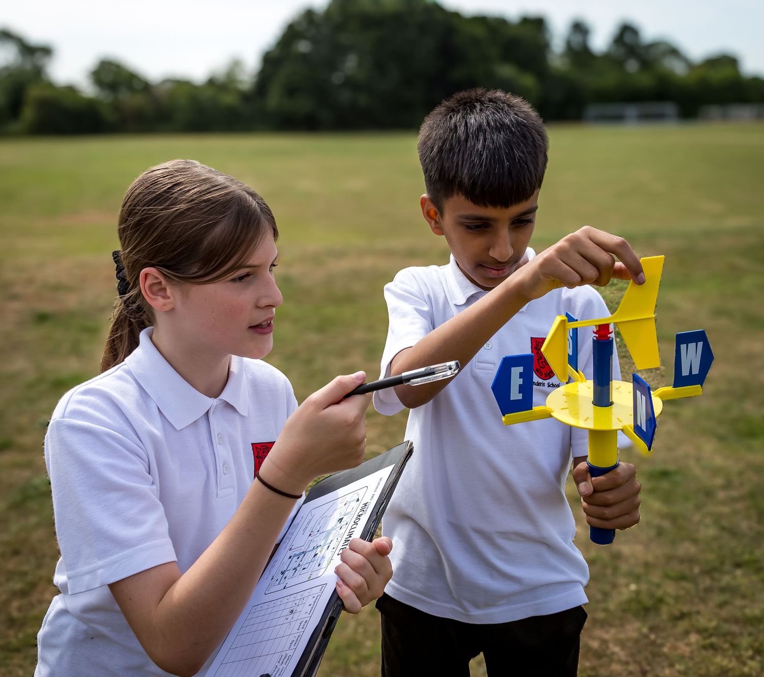 Two primary school children use a compass weather vane outdoors on a grass field during a science lesson.
