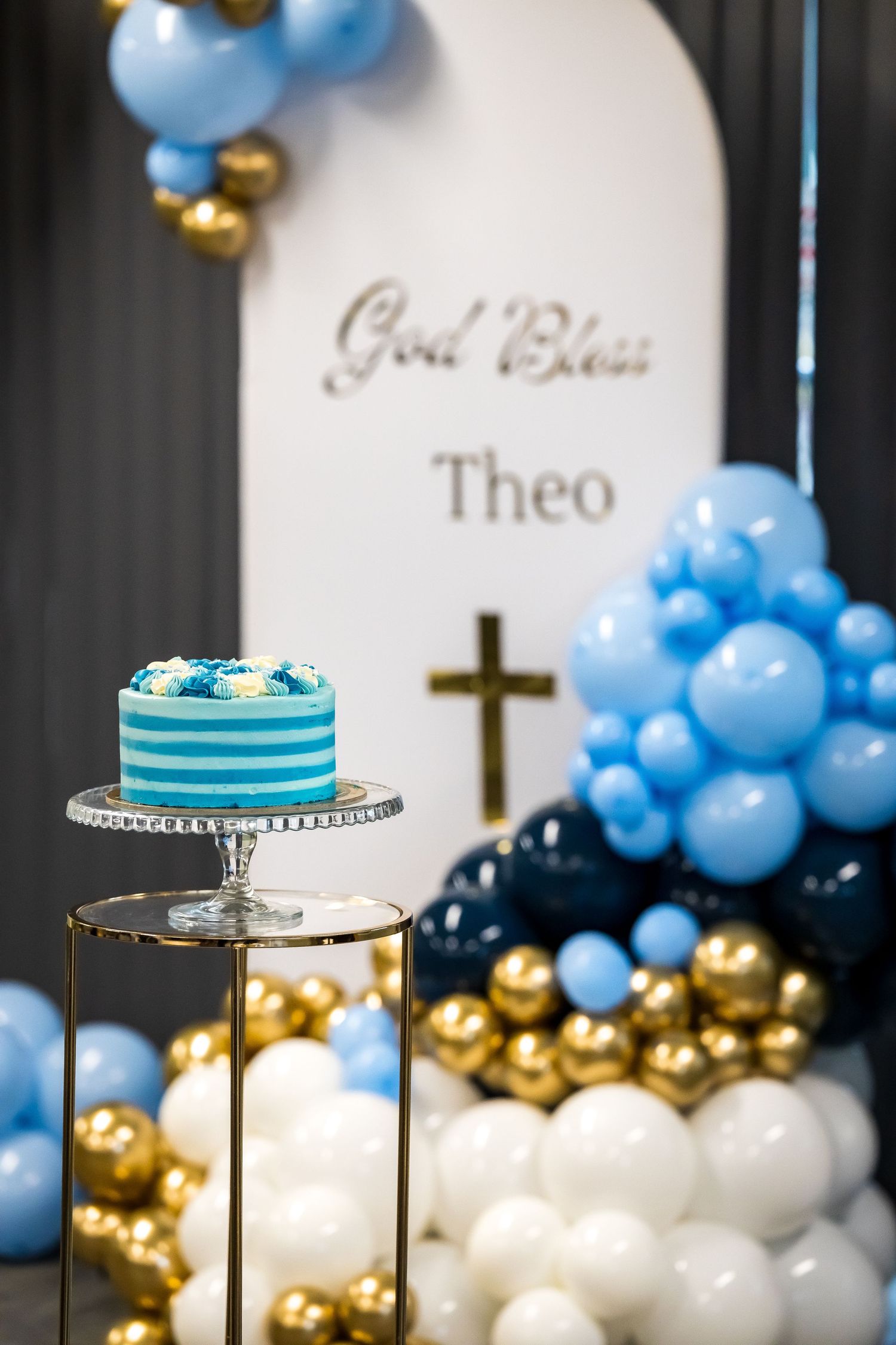 Blue and white christening cake on stand with God Bless Theo balloon arch and cross decoration.