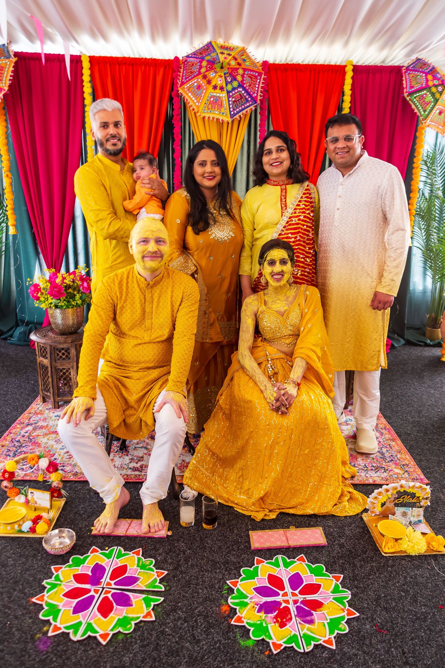 Indian family group photo in yellow outfits at a traditional Haldi ceremony with rangoli decorations.