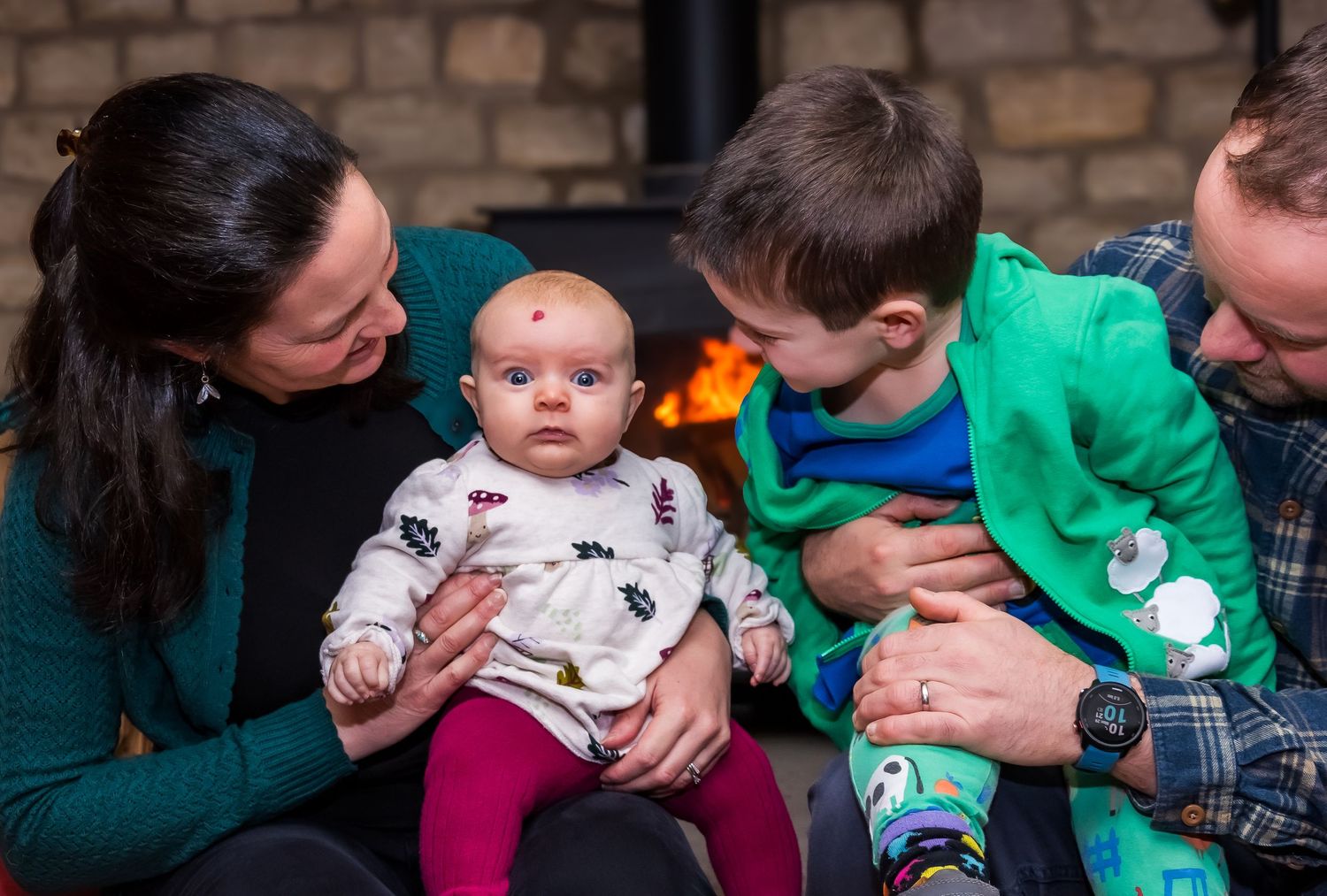A family of four with two parents, a toddler boy, and a baby sitting together near a fireplace.