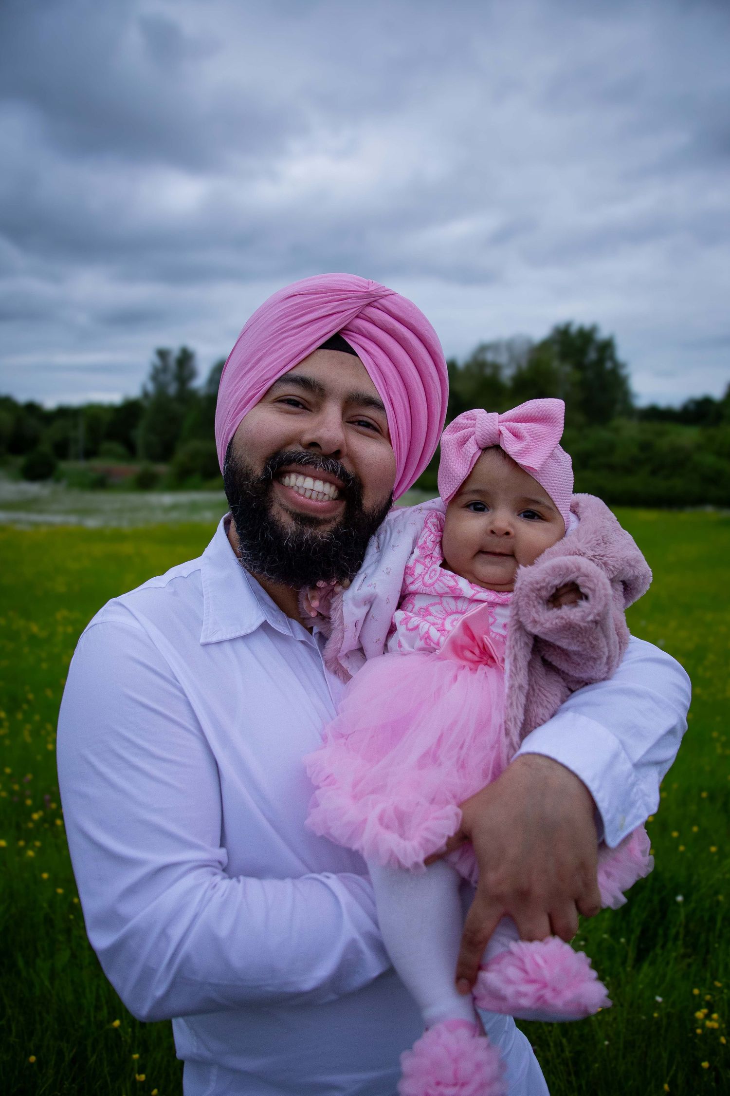 Smiling Sikh father in pink turban holding baby girl in pink outfit in green field.