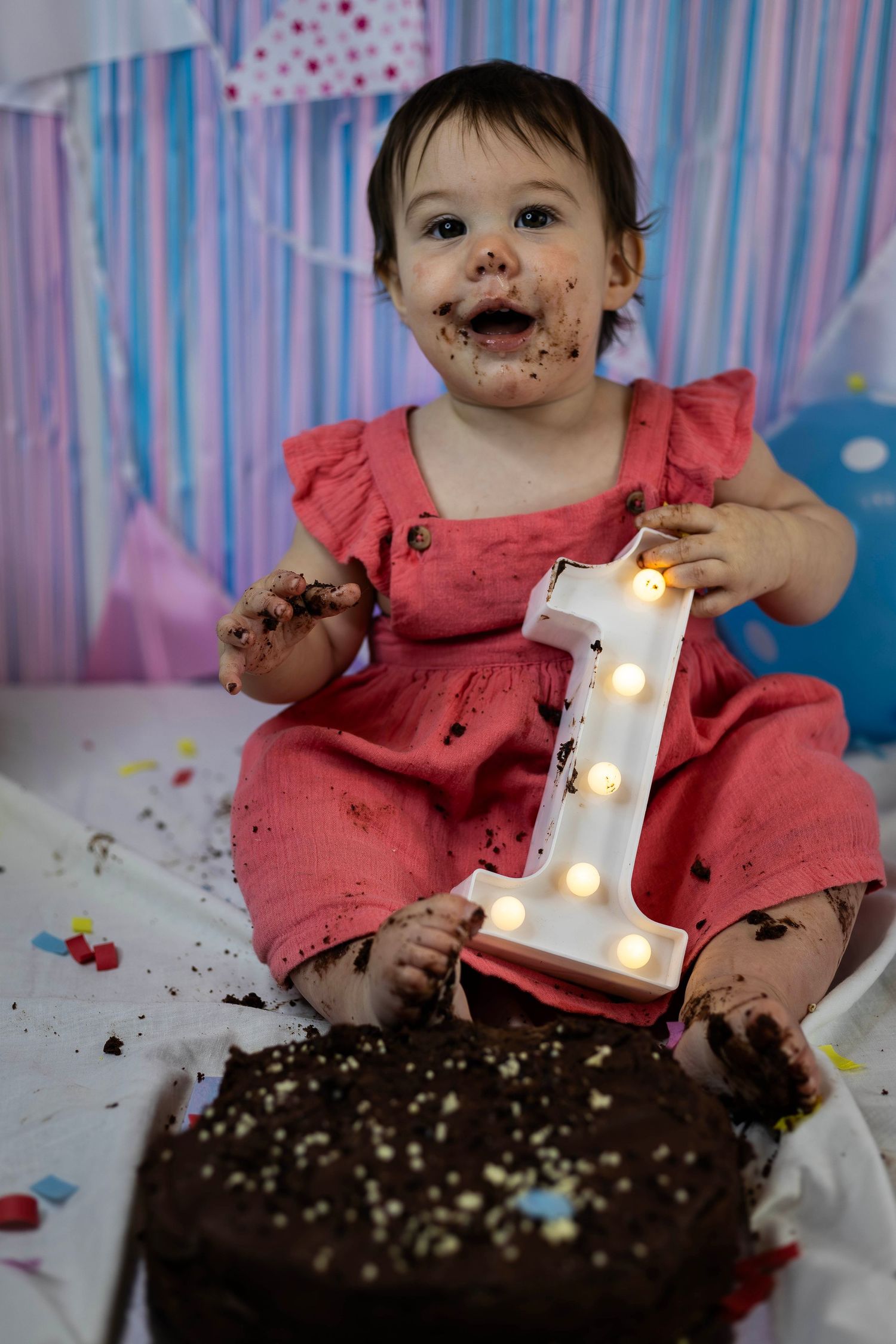 Baby girl in pink dress covered in chocolate cake celebrating her first birthday with illuminated number one.