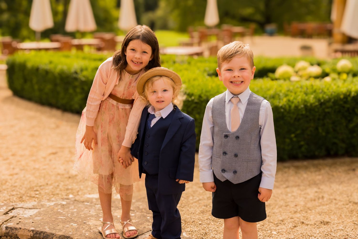 Three well-dressed children pose together outdoors at a wedding, smiling in formal attire on a sunny day.