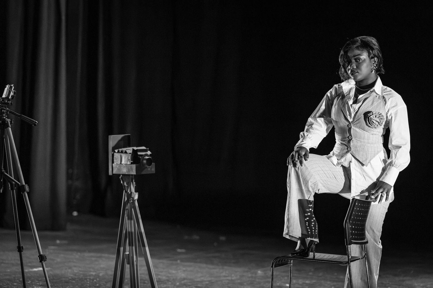 Black and white portrait of a person in white suit sitting on chair with vintage camera on stage.