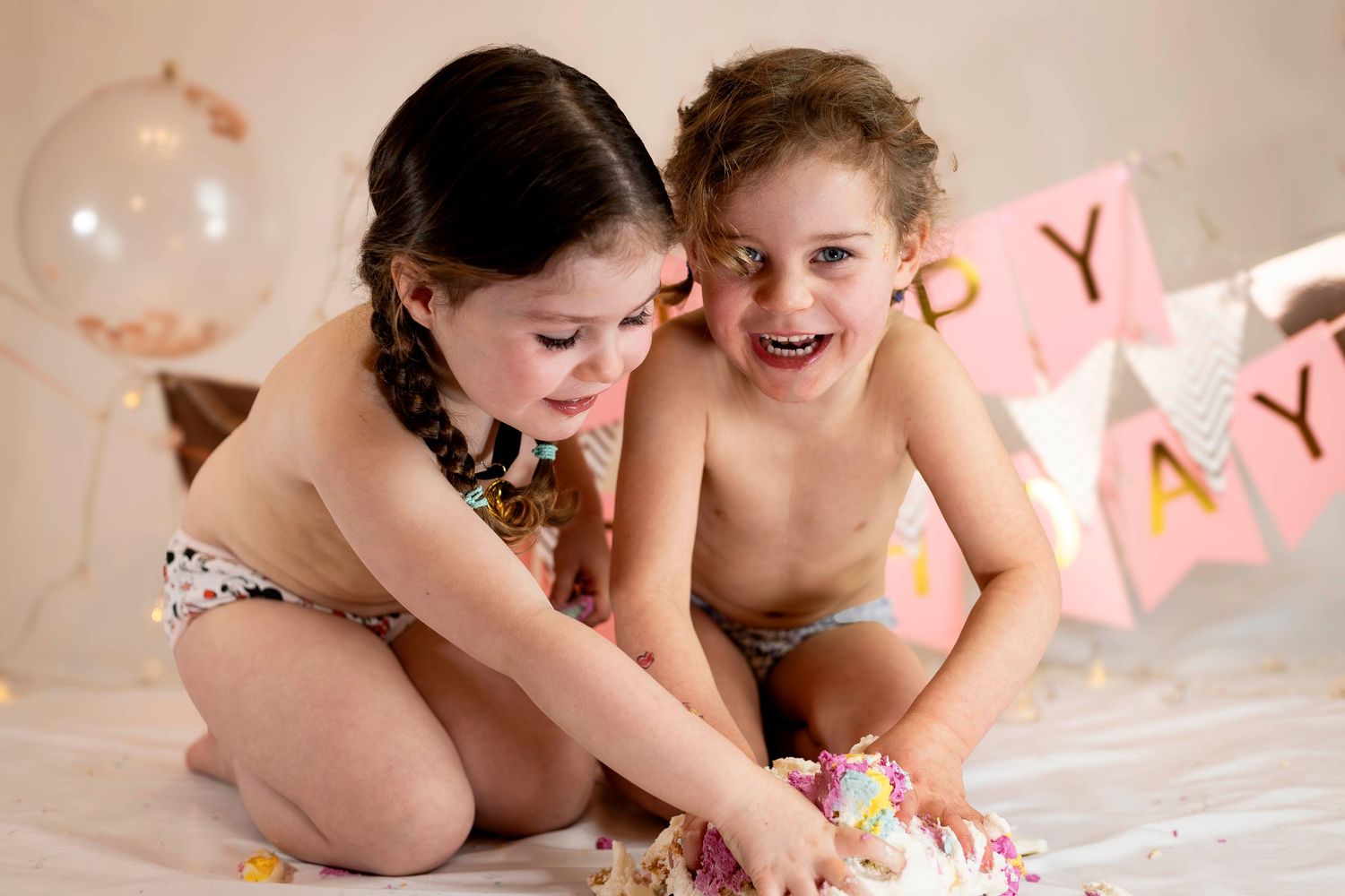 Two young girls laughing and smashing a birthday cake at a cake smash photoshoot.