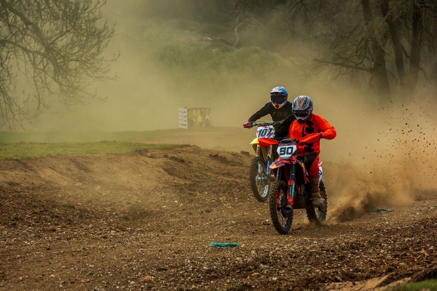 Two motocross riders race on a dirt track, kicking up dust and mud during an off-road motorcycle competition.