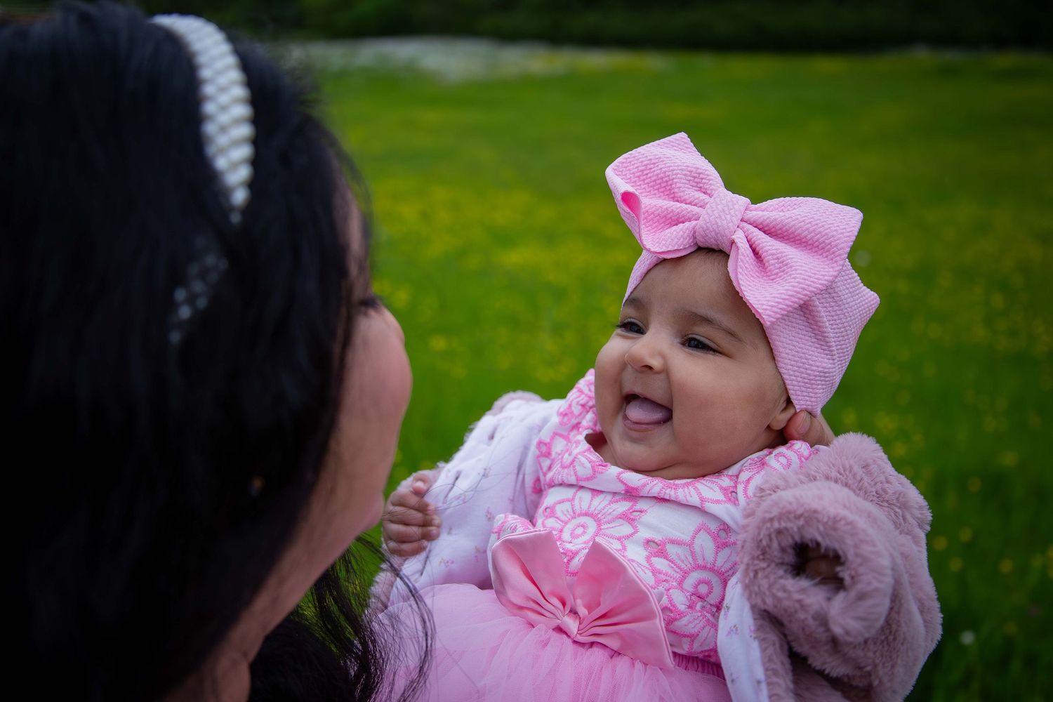 Adorable smiling baby in pink bow headband and dress being held by mother outdoors.