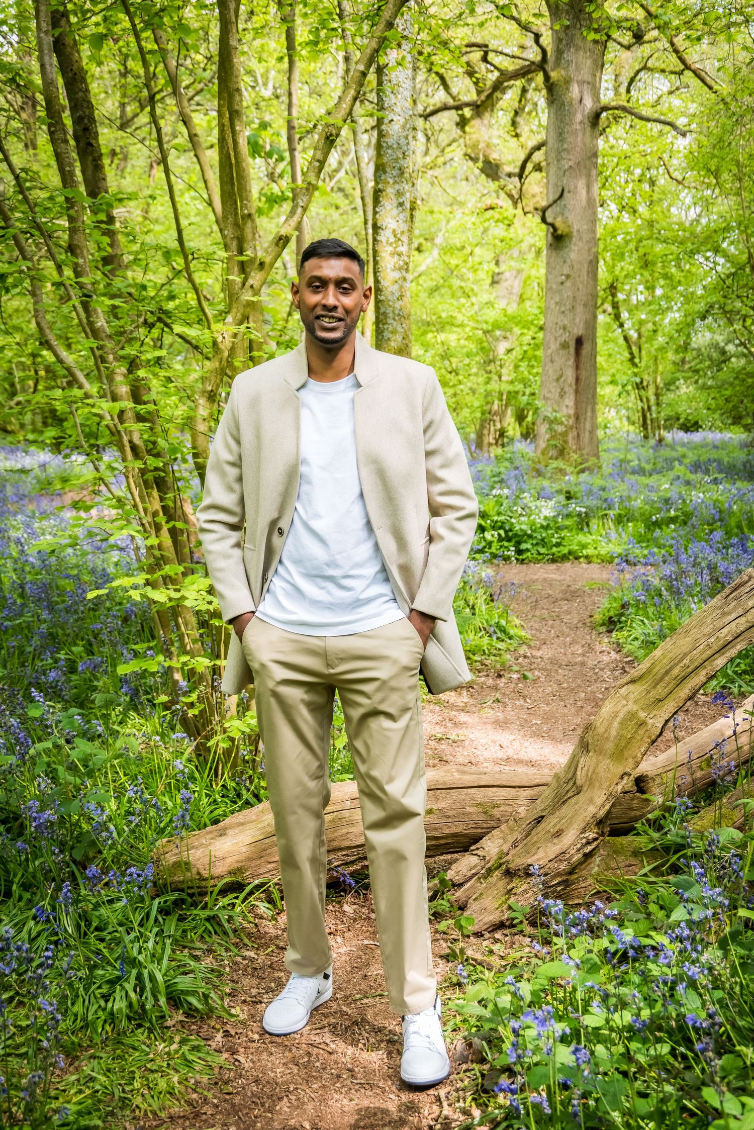 Smiling man in beige suit standing on a woodland path surrounded by bluebells and green trees.