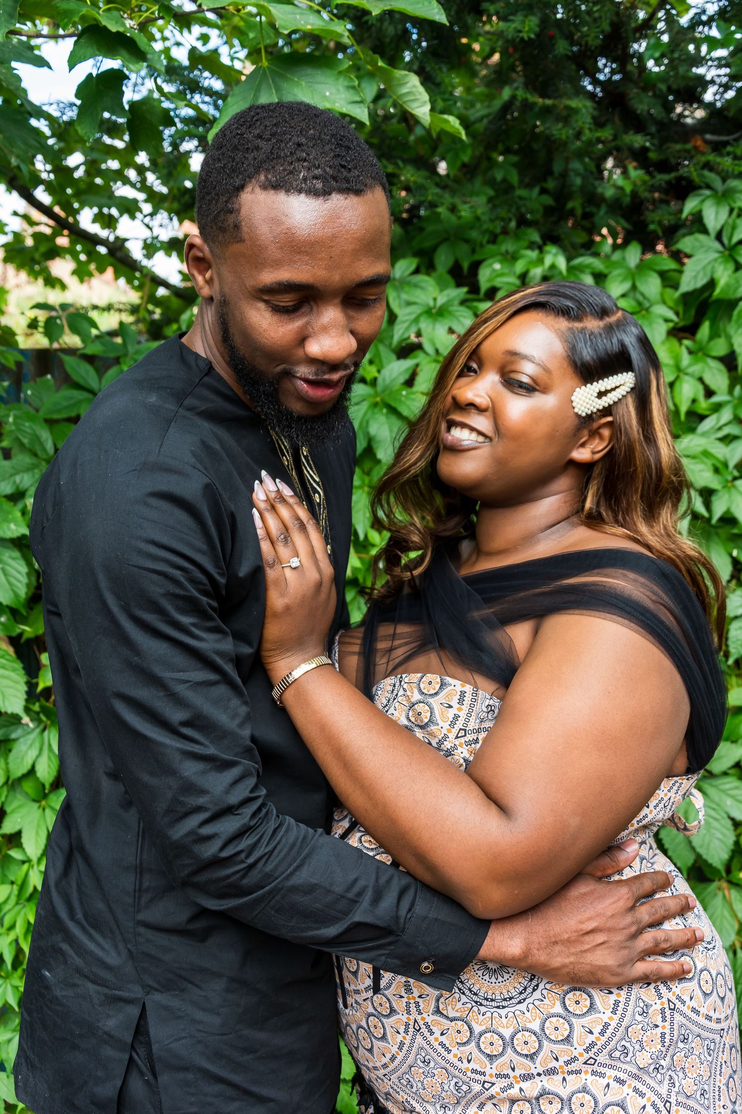 A couple embraces outdoors, smiling warmly, dressed in elegant attire with lush green foliage in the background.