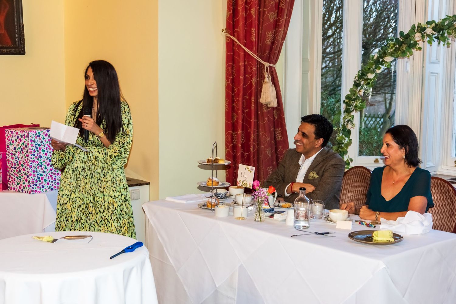 Woman speaking at podium during formal event with two guests seated at decorated table.