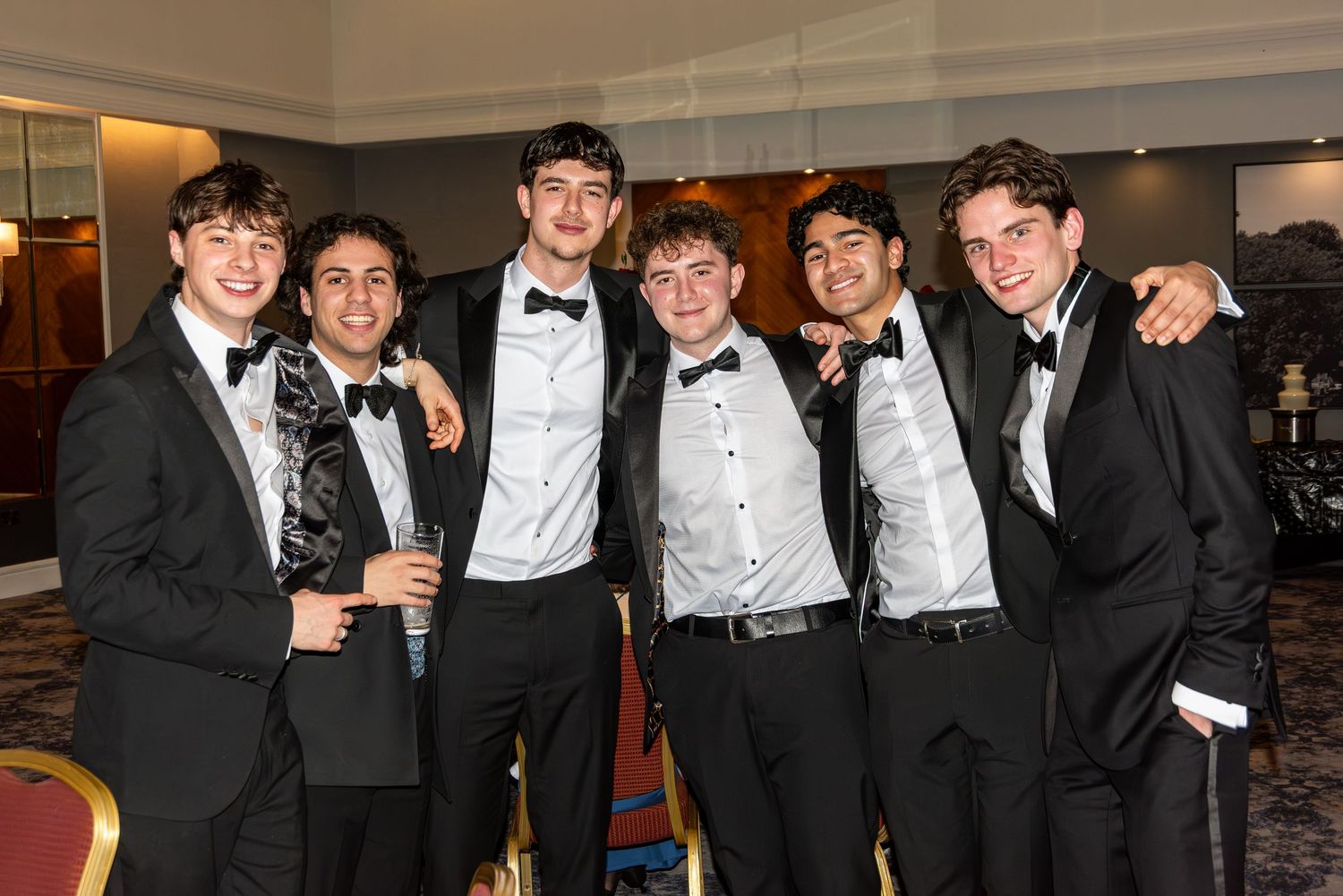 Six young men in black tuxedos and bow ties smiling together at a school ball.
