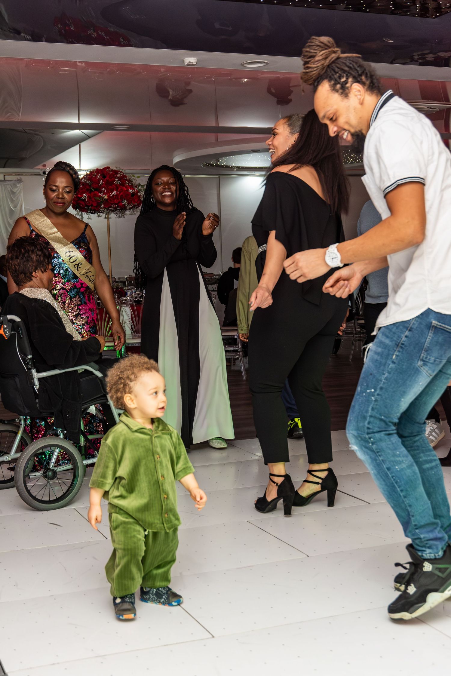 Toddler in green outfit dancing with adults at a lively indoor party.