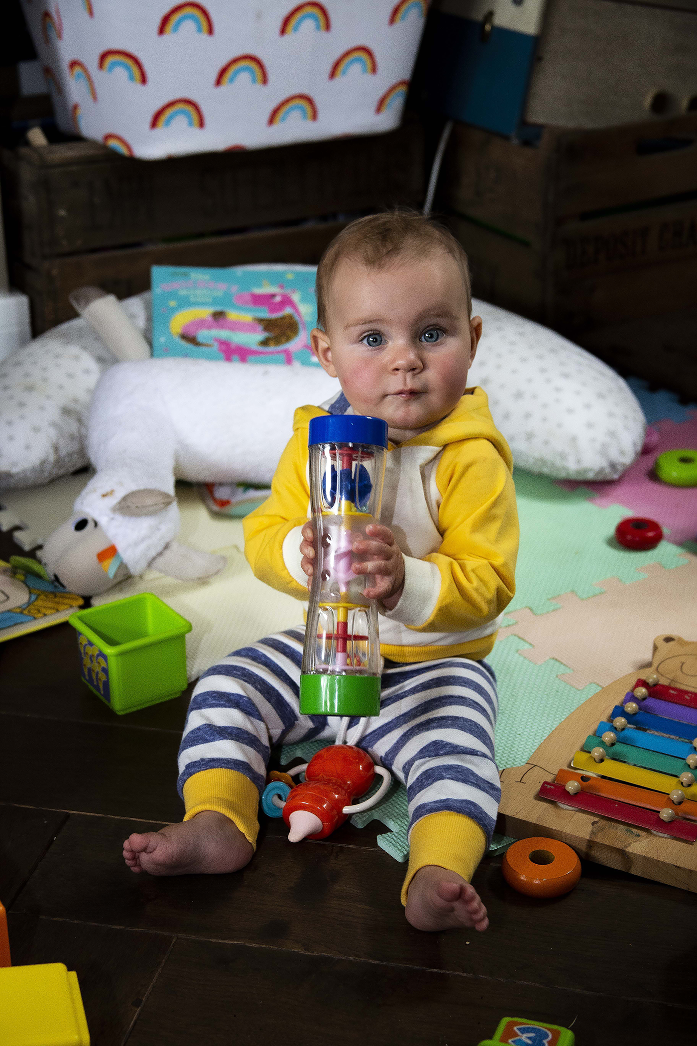 Baby boy in yellow hoodie sitting on floor surrounded by colorful toys, holding a rain stick toy.