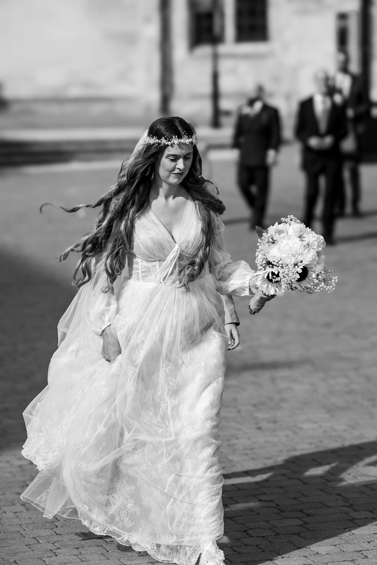 A bride with long flowing hair and a floral crown strides gracefully in a boho wedding gown, black and white.
