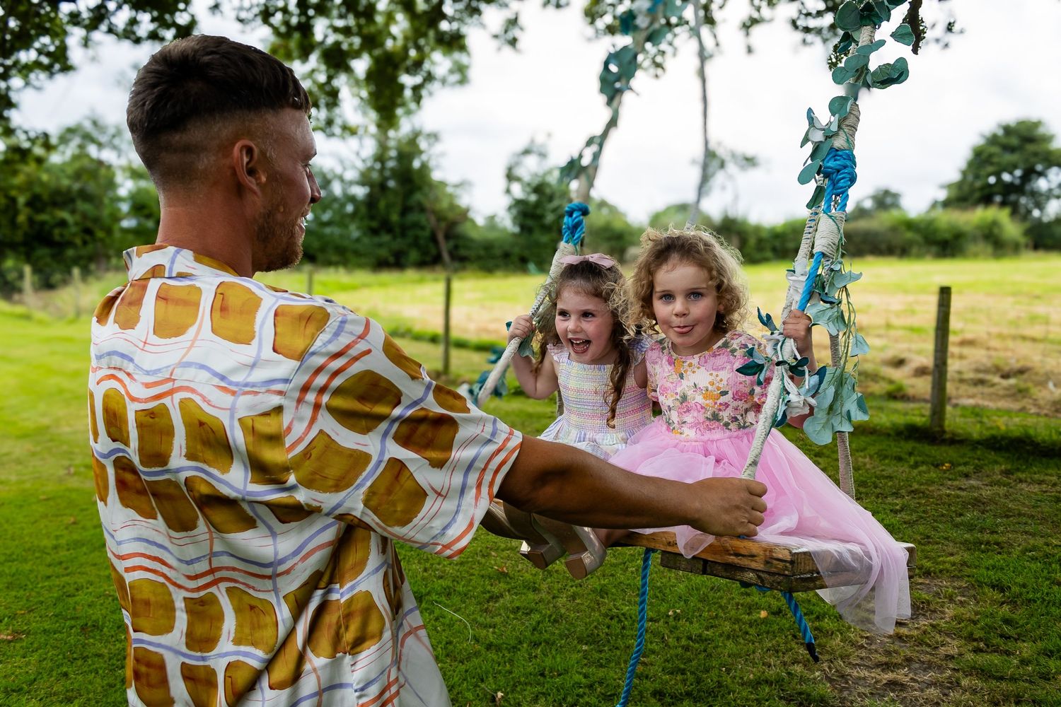 Man pushing two young girls on a decorative rope swing in a lush green garden.