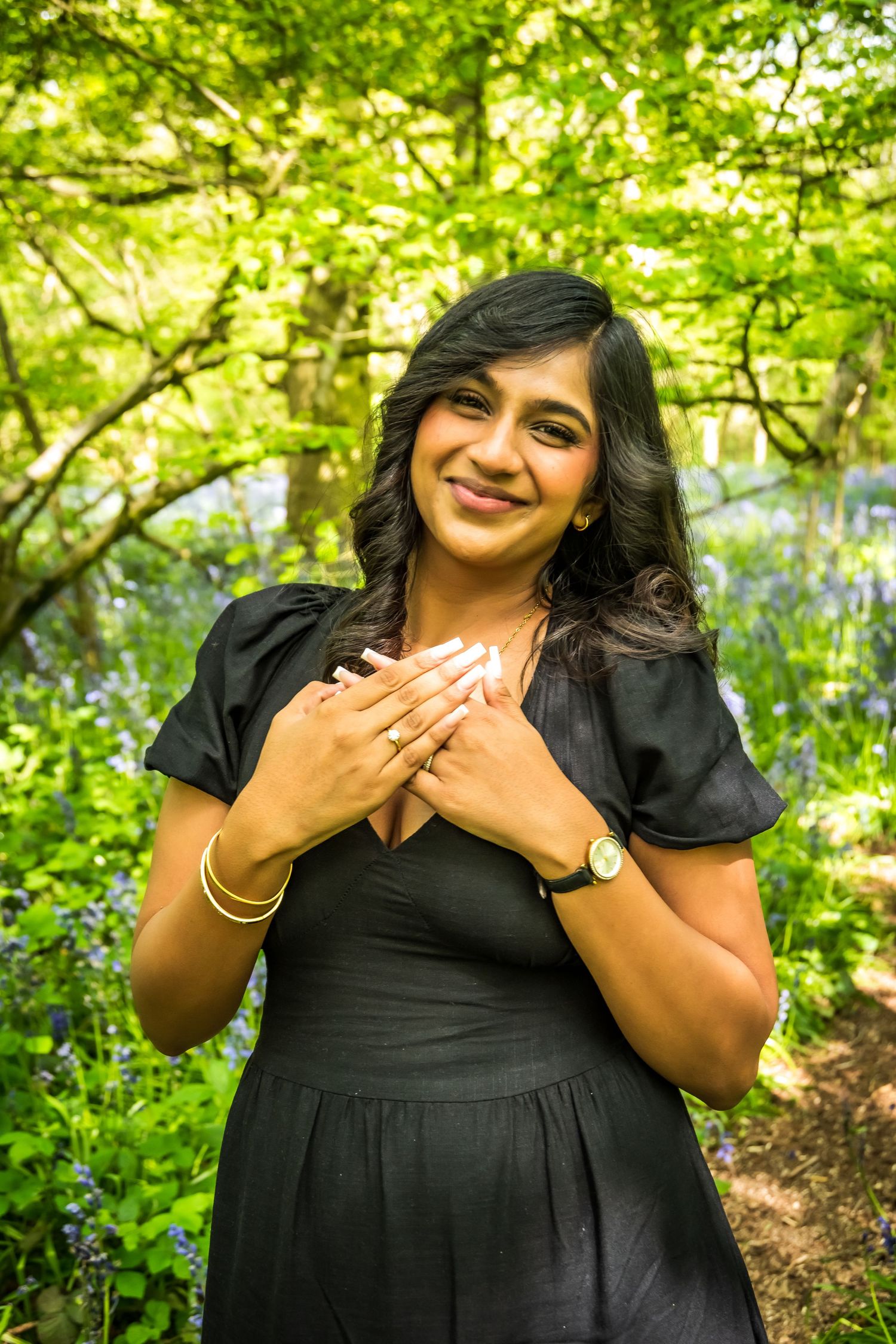 A smiling woman in a black dress poses with hands on chest in a lush green forest with bluebells.