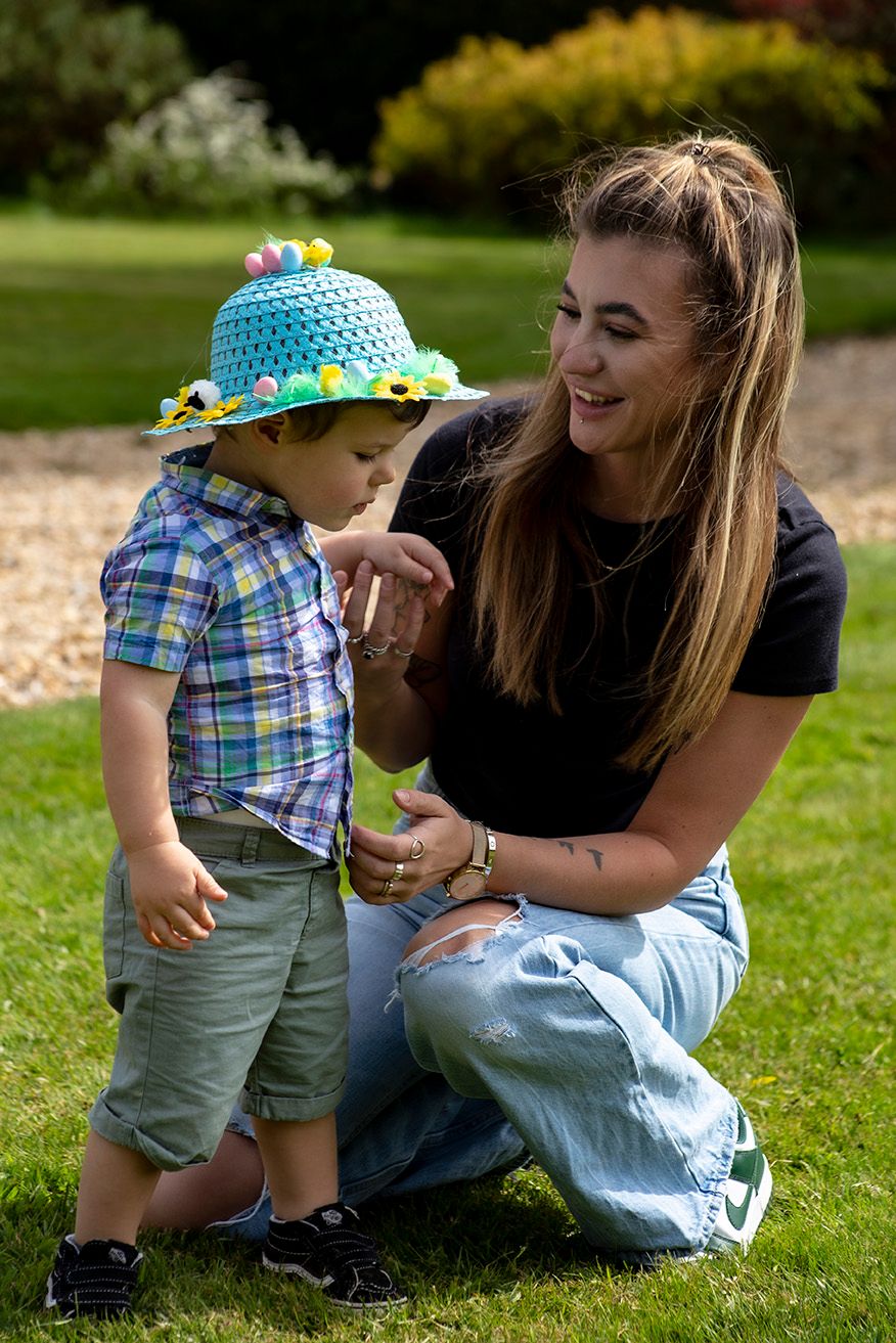 Young woman kneeling beside toddler boy wearing colorful Easter hat in sunny garden setting.