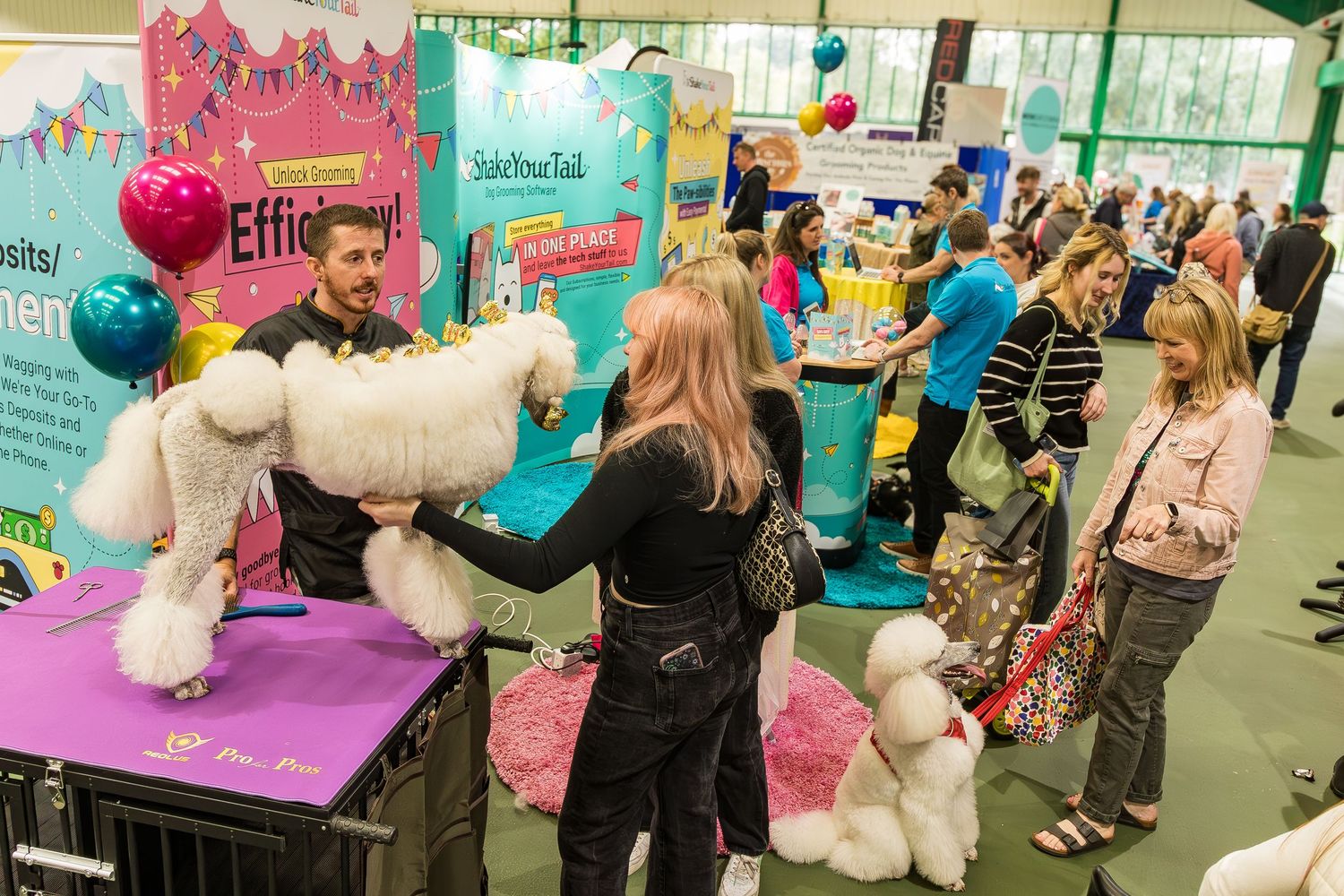 A lively pet expo with a groomer styling a white poodle while visitors browse vendor booths and dogs.