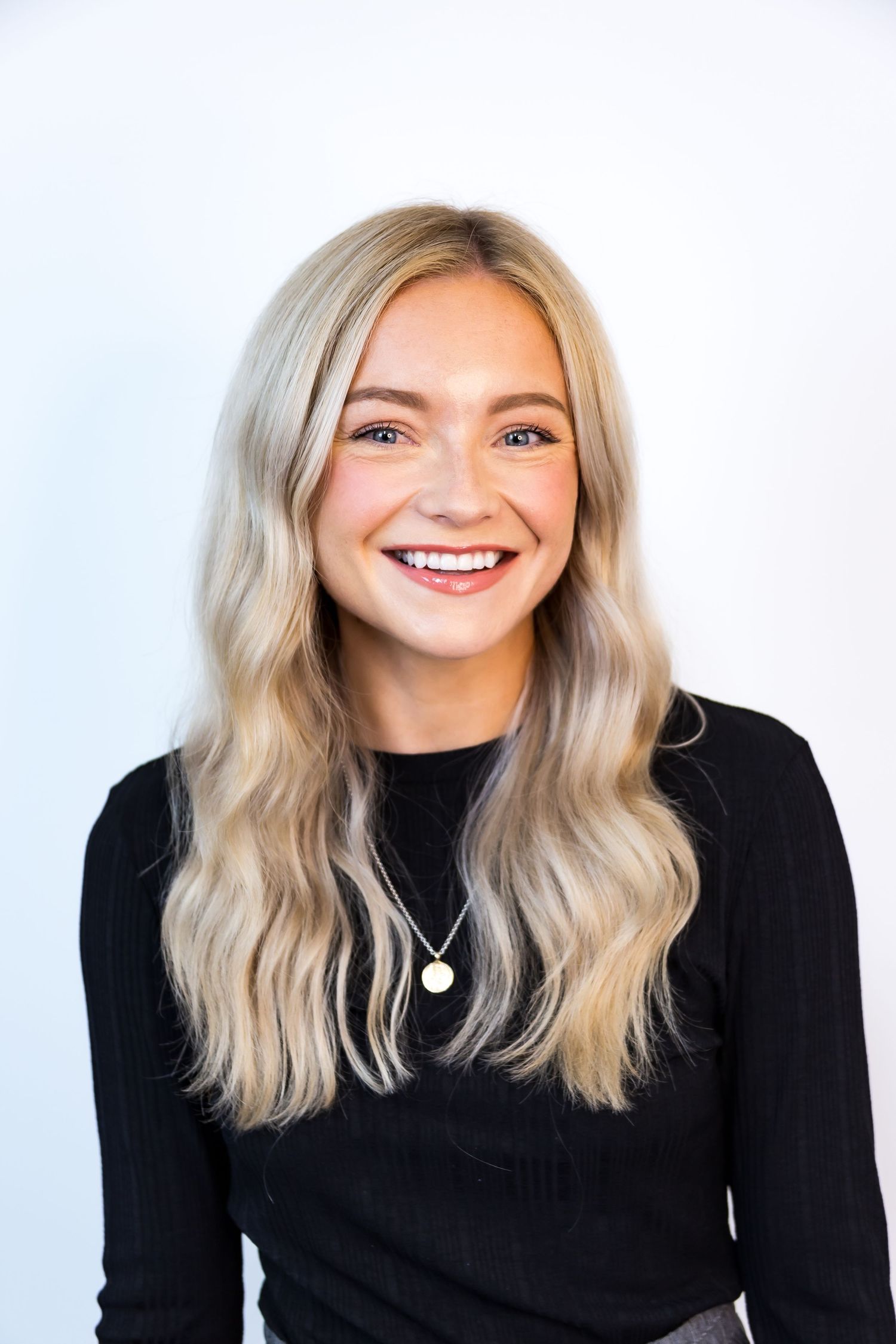 A smiling young blonde woman in a black top poses for a professional headshot against a white background.