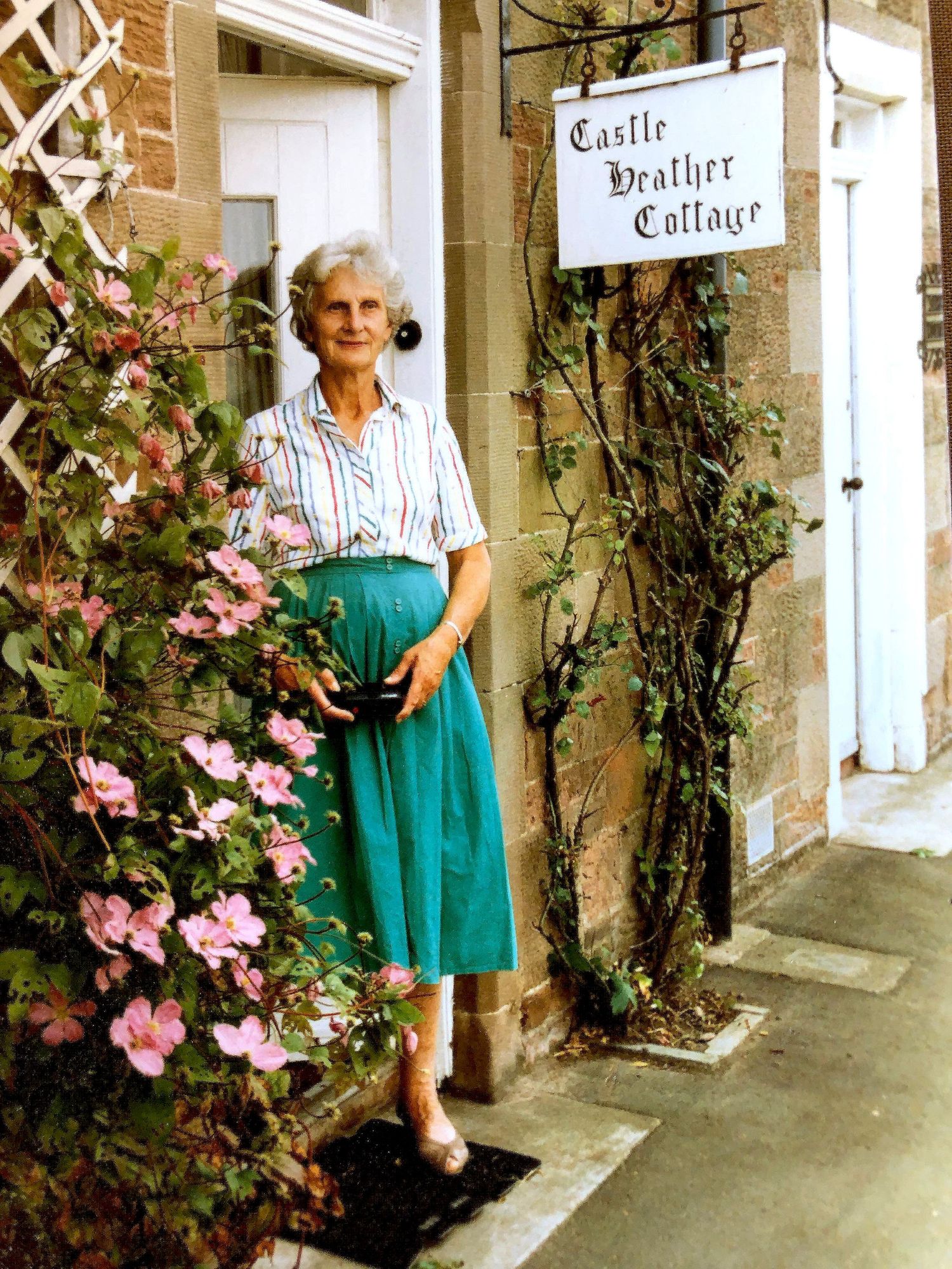 Elderly woman in teal skirt standing outside Castle Heather Cottage, surrounded by pink climbing roses.