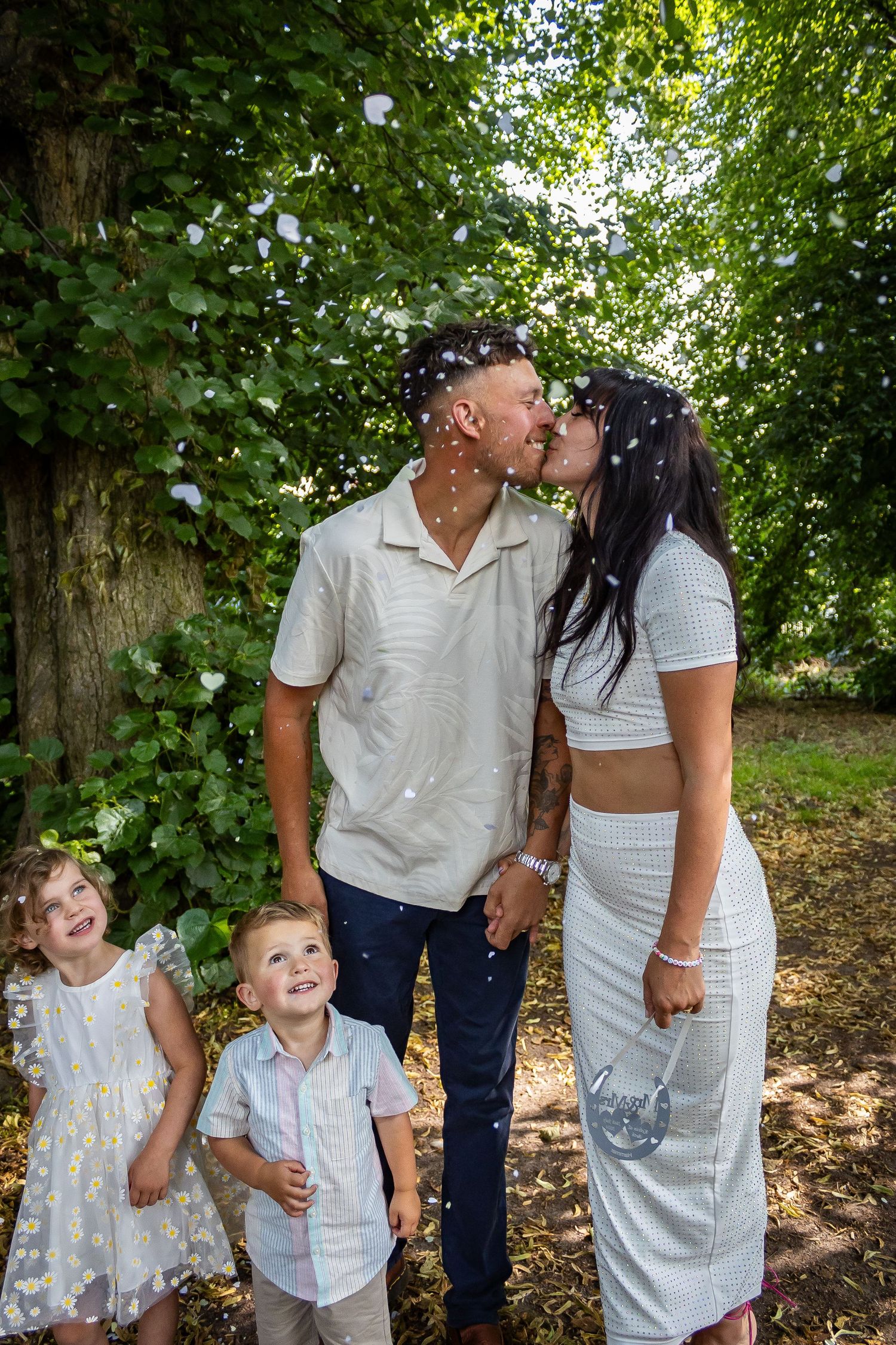 Couple kissing outdoors as confetti falls while two young children laugh and smile beneath lush green trees.
