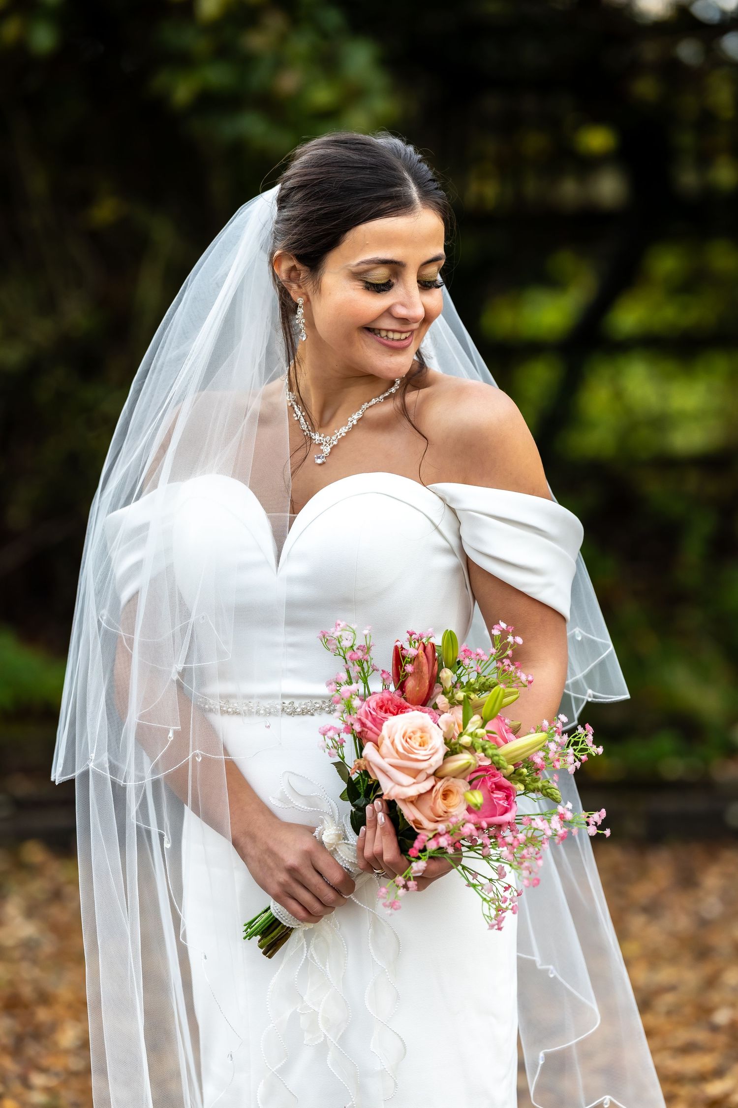 Beautiful bride in off-shoulder white gown holding a pink floral bouquet with veil.
