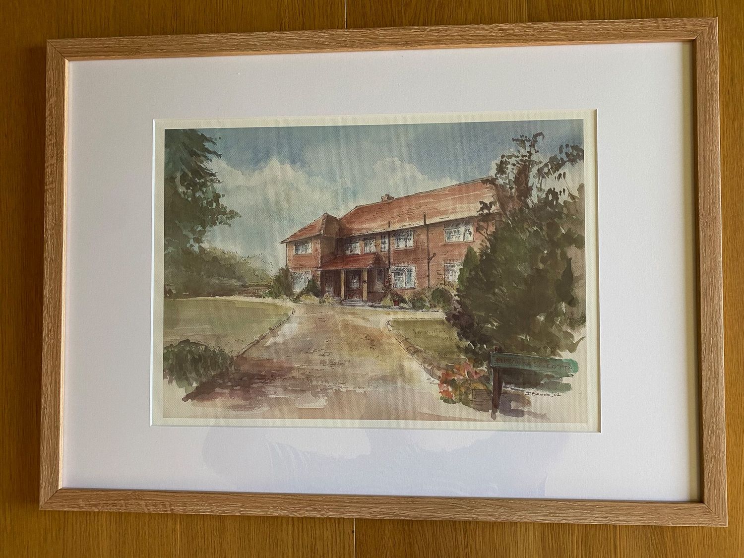 Framed watercolor painting of a red brick building surrounded by trees, displayed in a wooden frame with white matting.