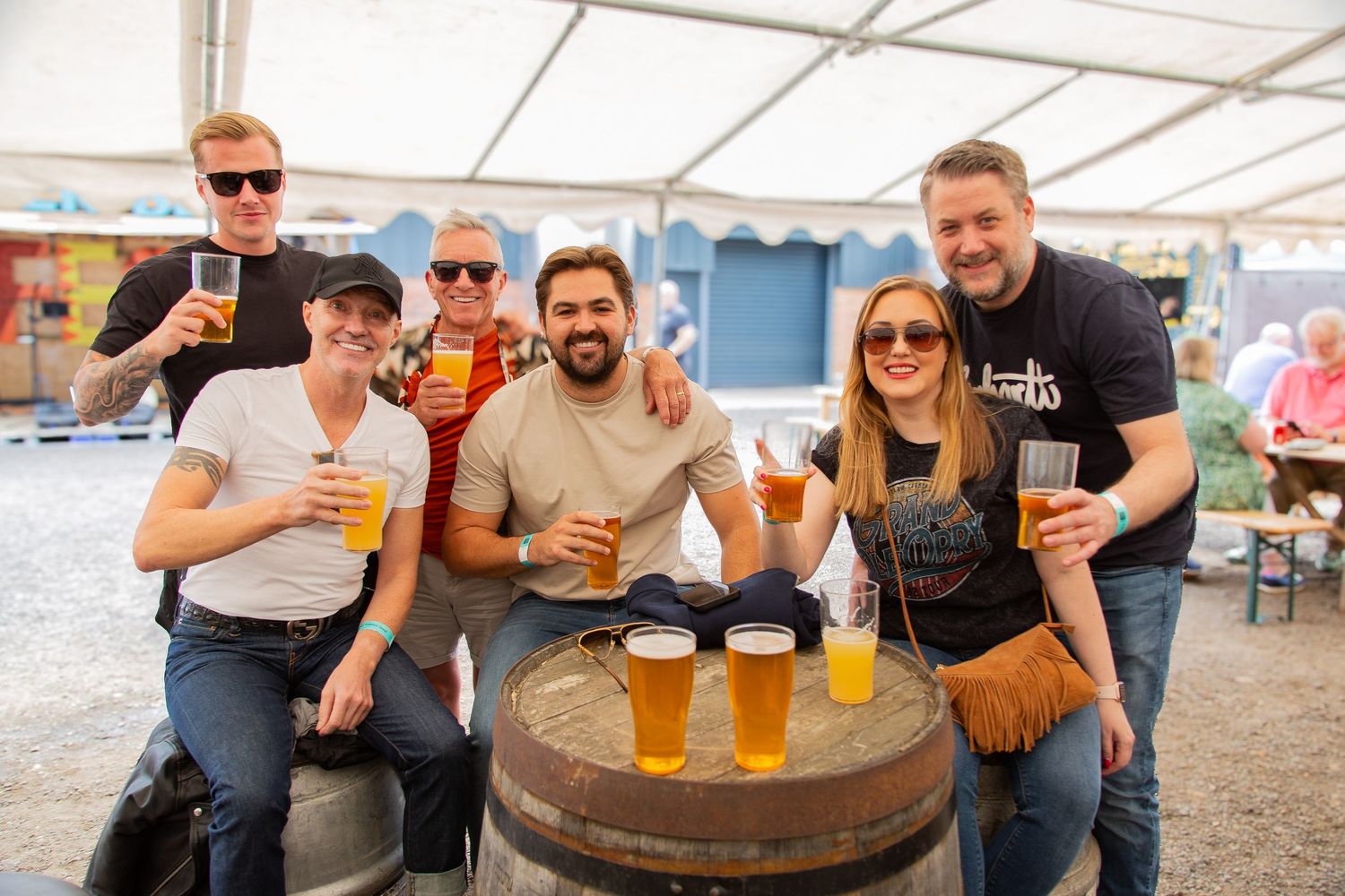 Group of friends smiling and posing with pints of beer around a barrel at an outdoor beer festival.
