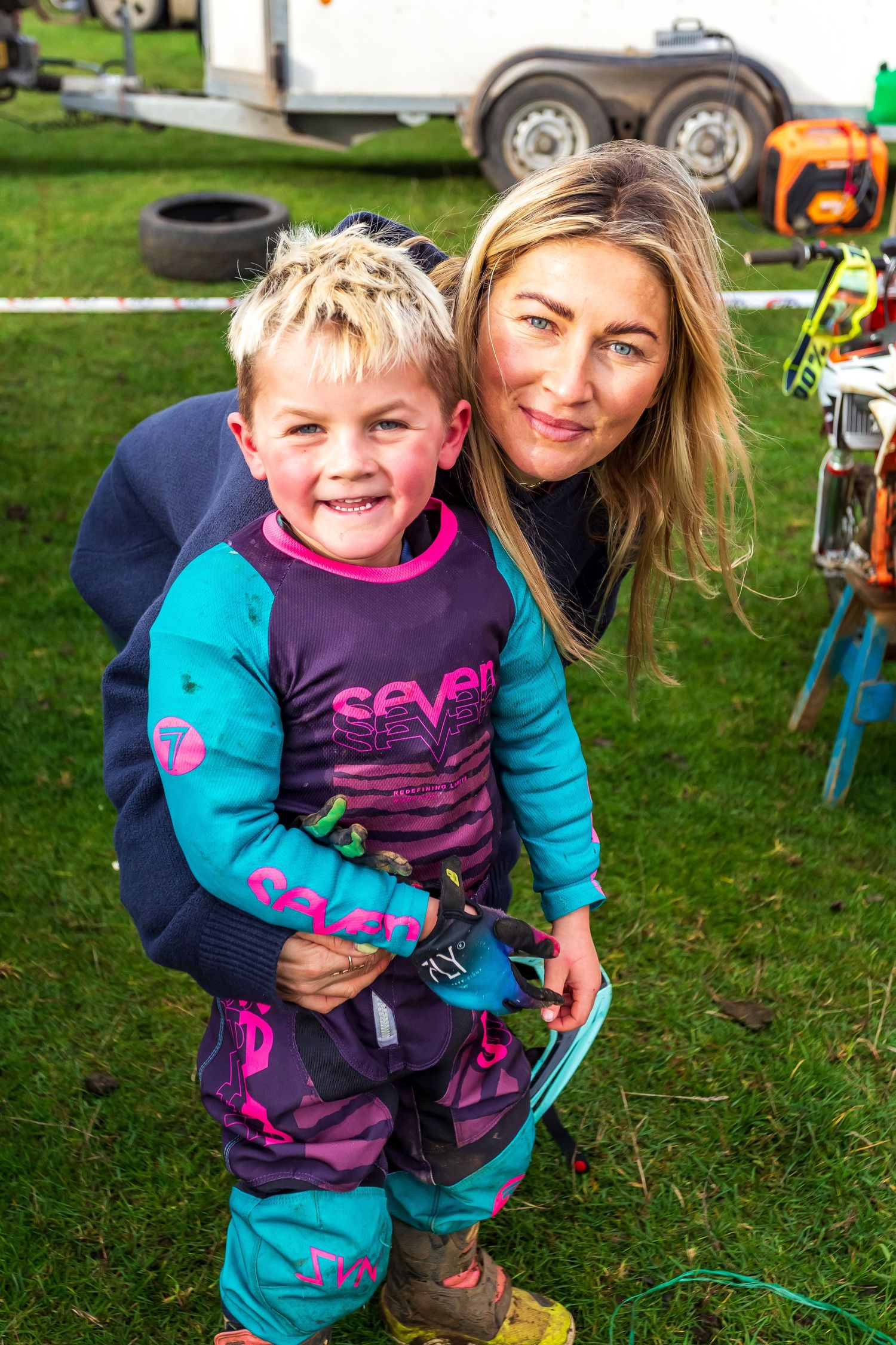 Young boy in motocross gear smiles with a woman outdoors near trailers and motorcycles at a racing event.