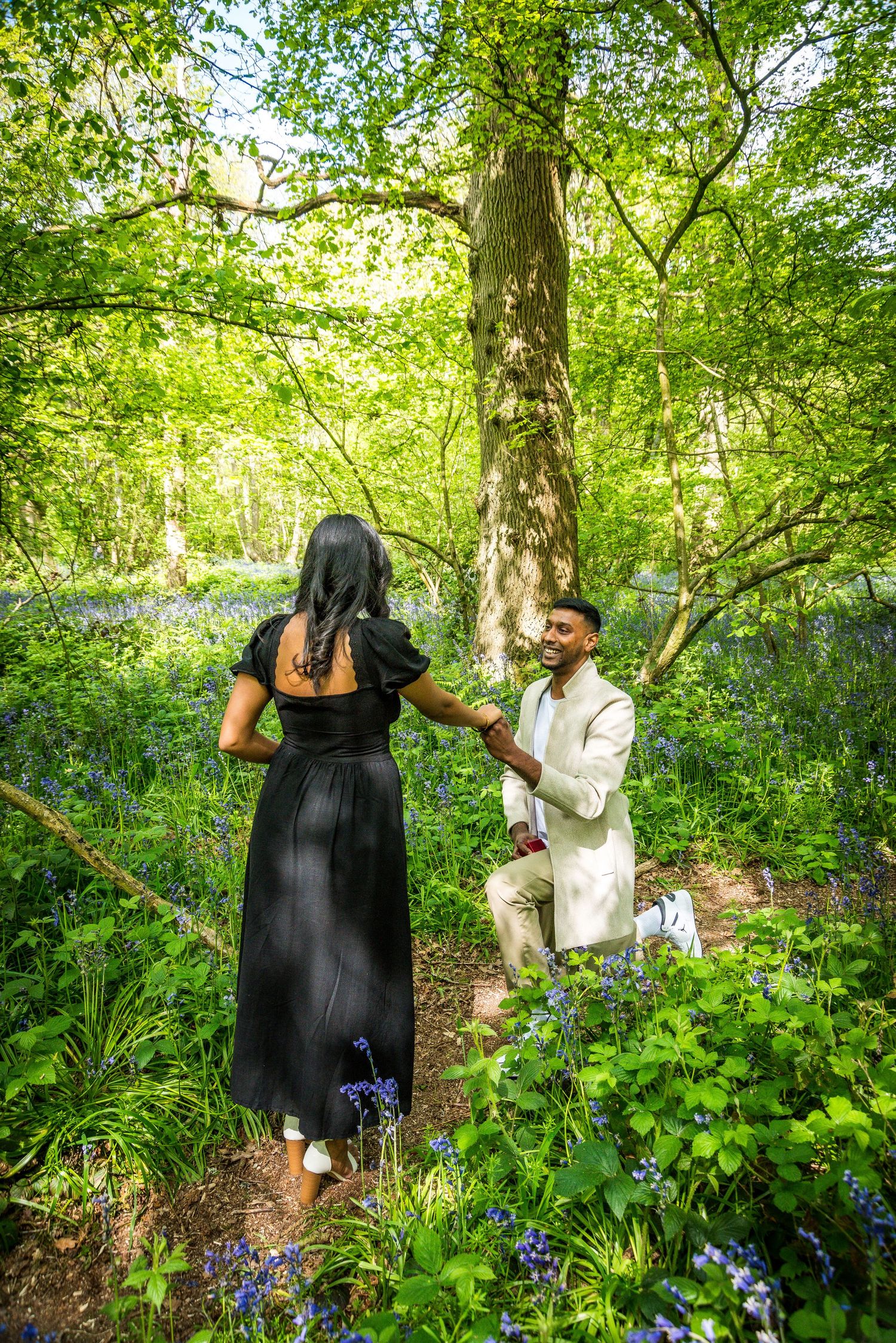 Man kneeling on one knee proposing to woman in black dress among bluebells in a lush green woodland.