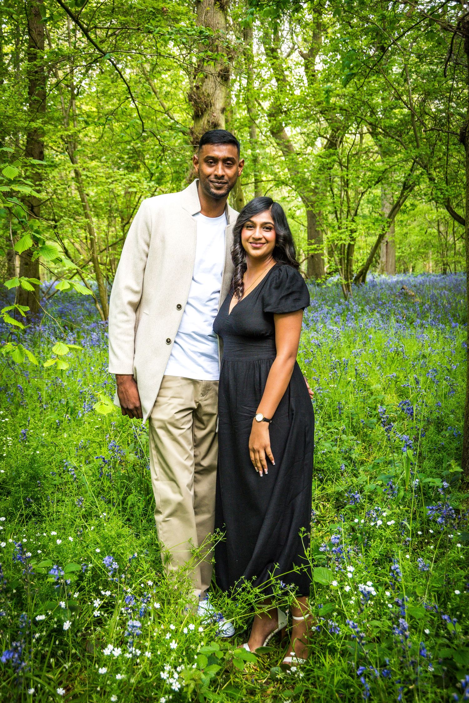 Couple posing together in a bluebell woodland, man in beige jacket and woman in black dress.