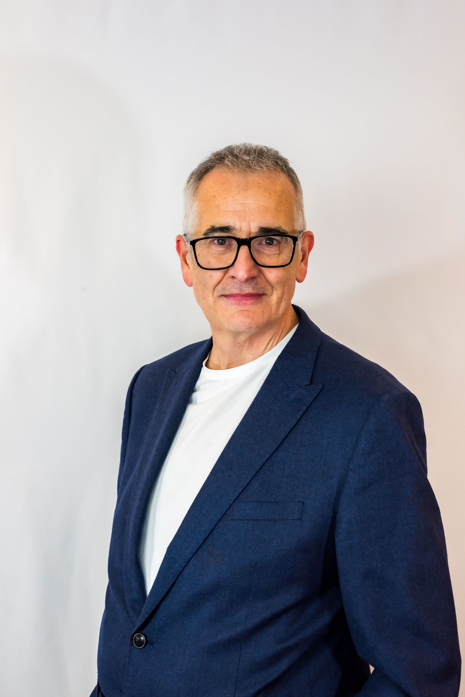 Middle-aged man with glasses wearing blue blazer and white t-shirt against light background.