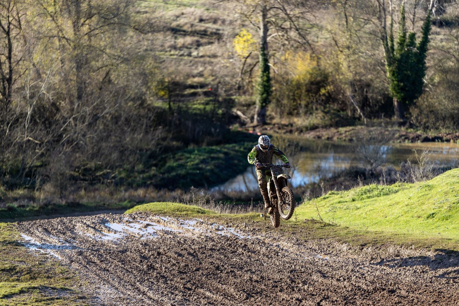 Motocross rider navigating a muddy off-road dirt track surrounded by trees and a pond in a racing event in the UK.