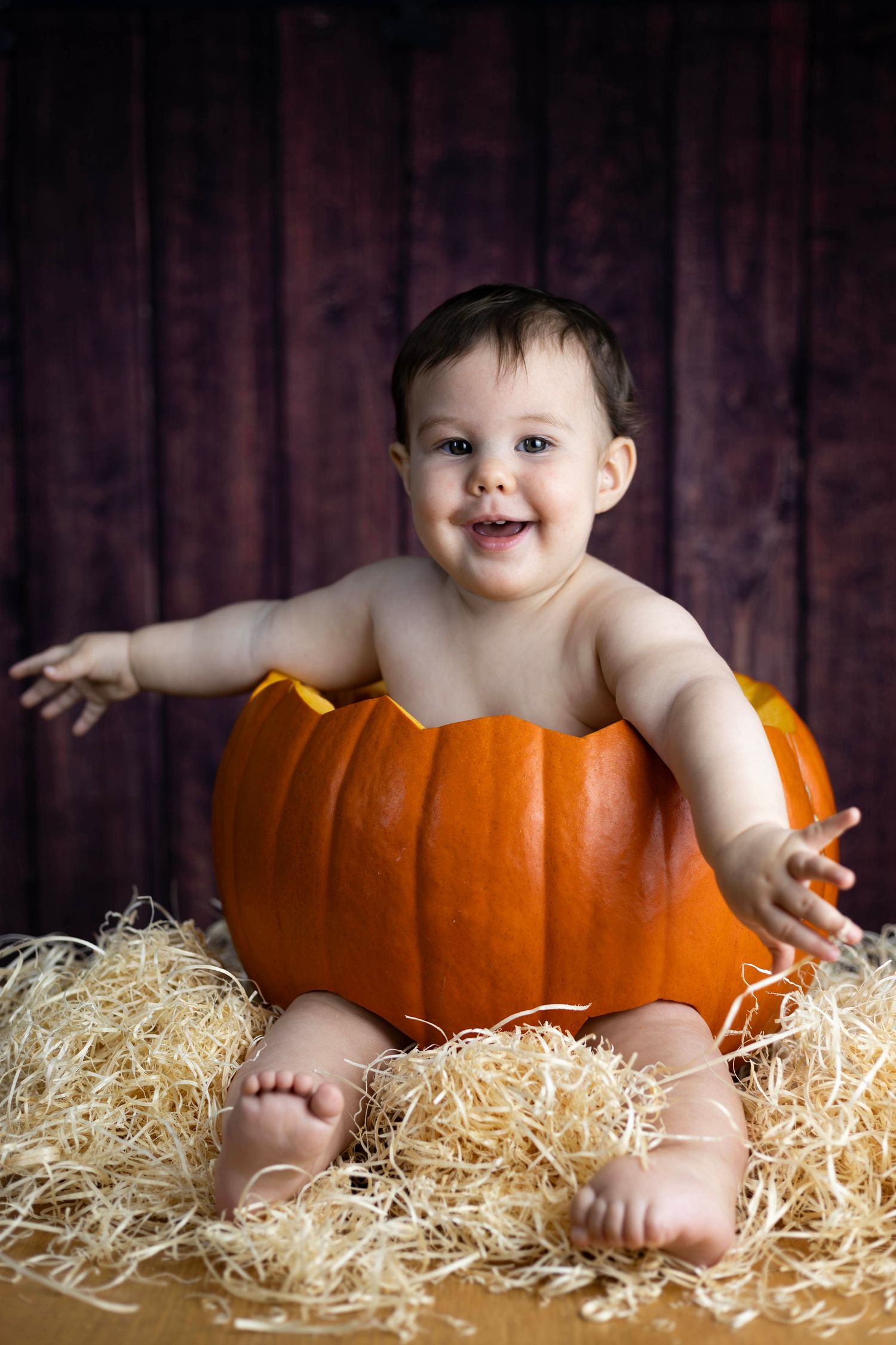 Cheerful baby sitting inside a large orange pumpkin on straw with arms outstretched, smiling.