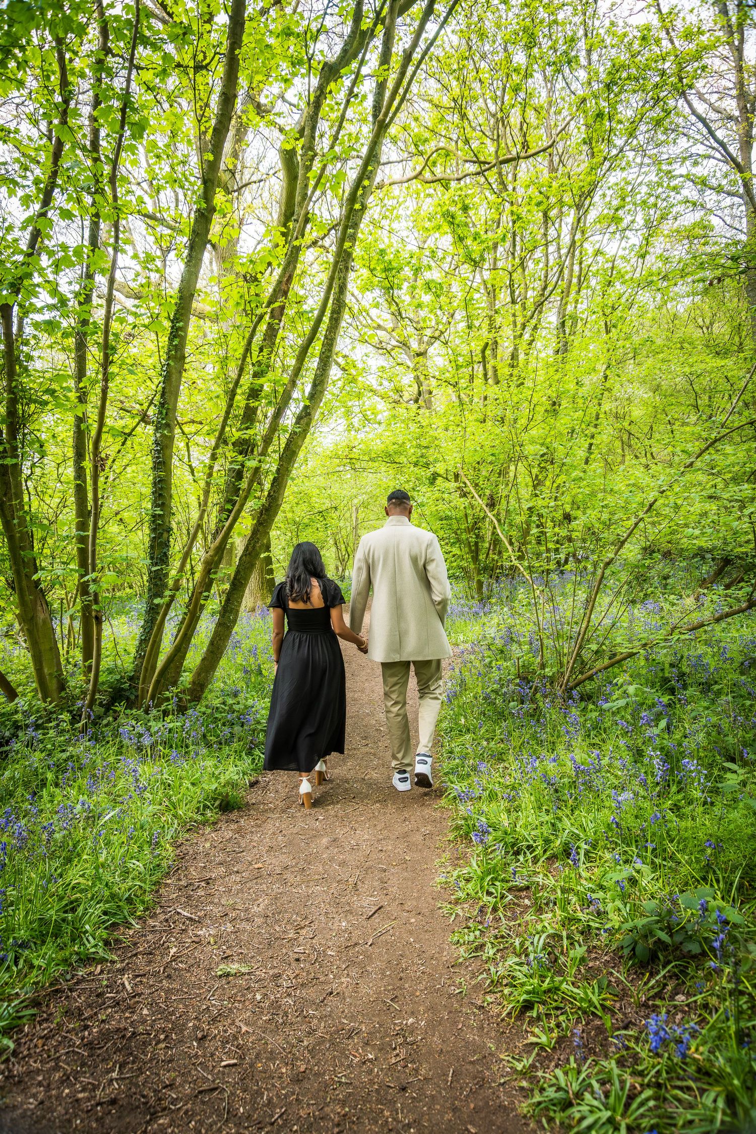 Couple walking hand in hand along a woodland path surrounded by bluebell flowers and green trees.