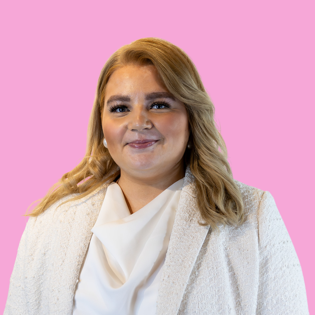 Professional headshot of a smiling blonde woman in a white blazer against a pink background.