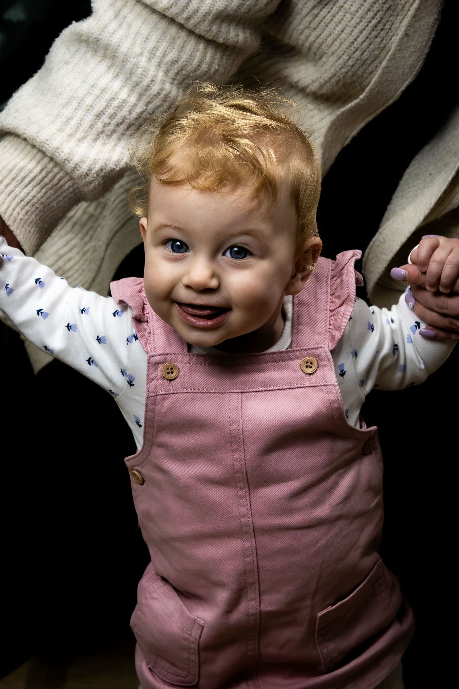 Smiling baby girl in pink dungarees stands with arms raised, held by adult hands against dark background.