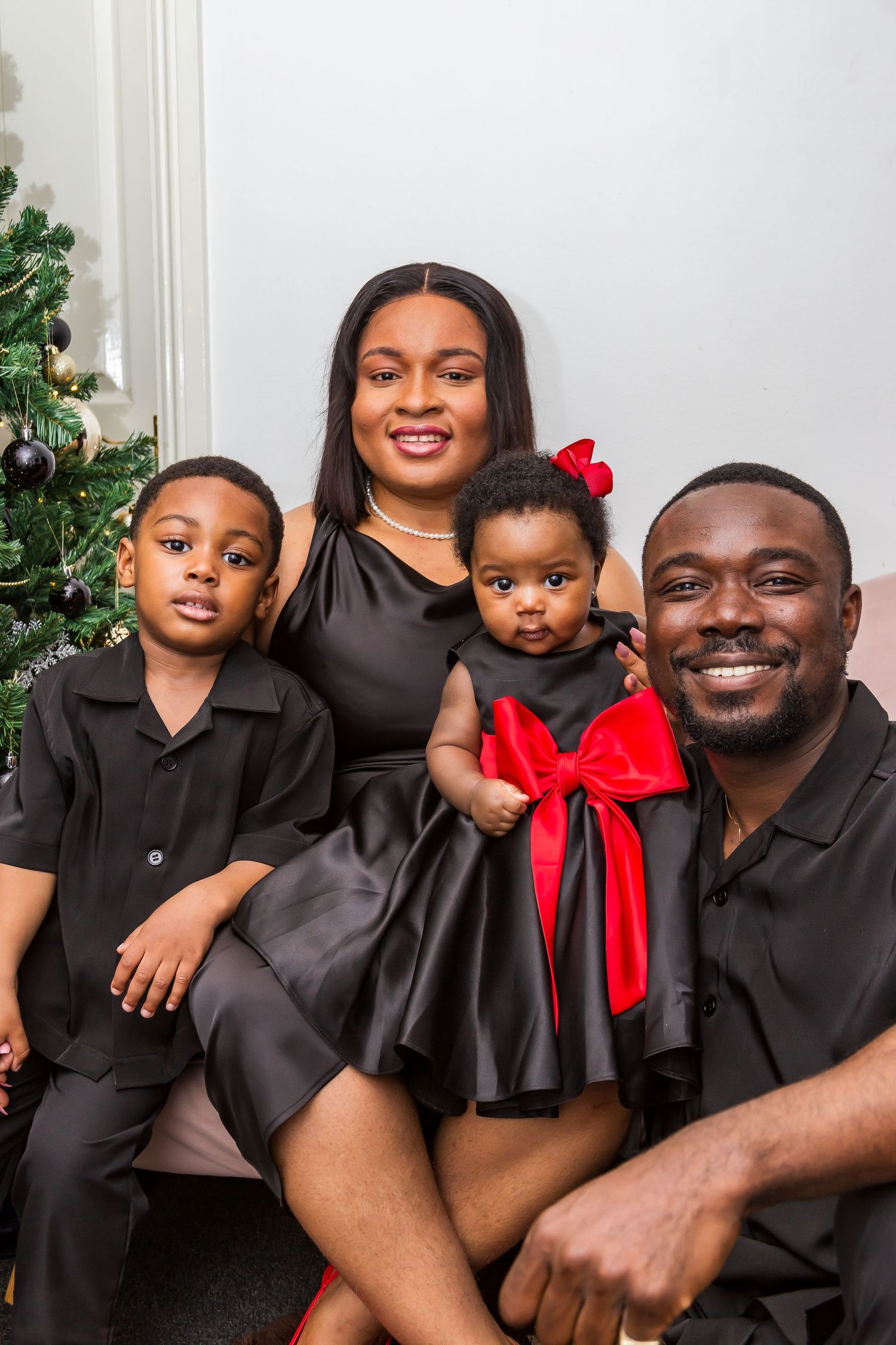 Festive family Christmas portrait with parents and two children dressed in coordinating black outfits near a tree.