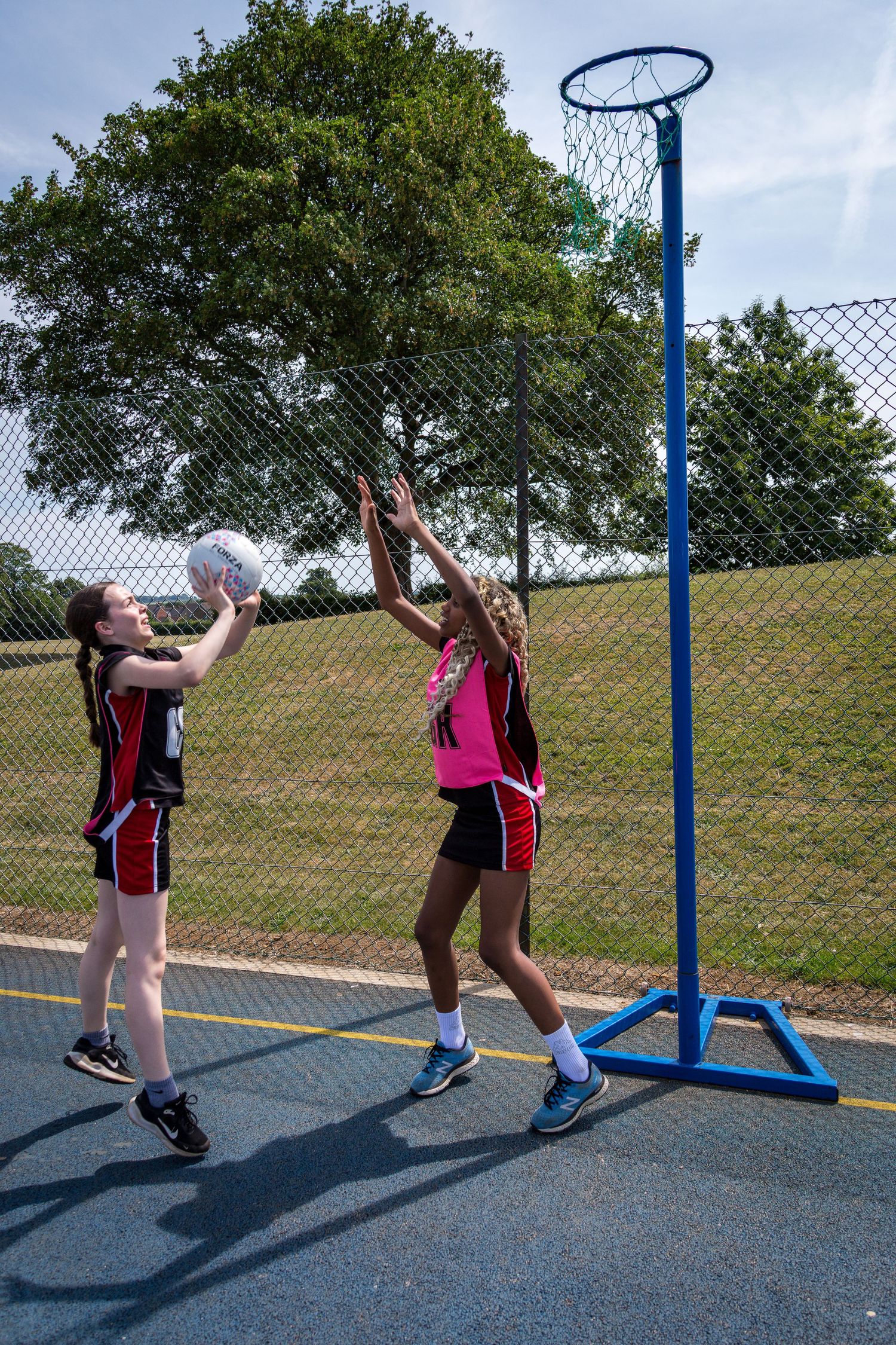 Two girls playing netball, jumping for the ball near the post on an outdoor court in a school in the UK.