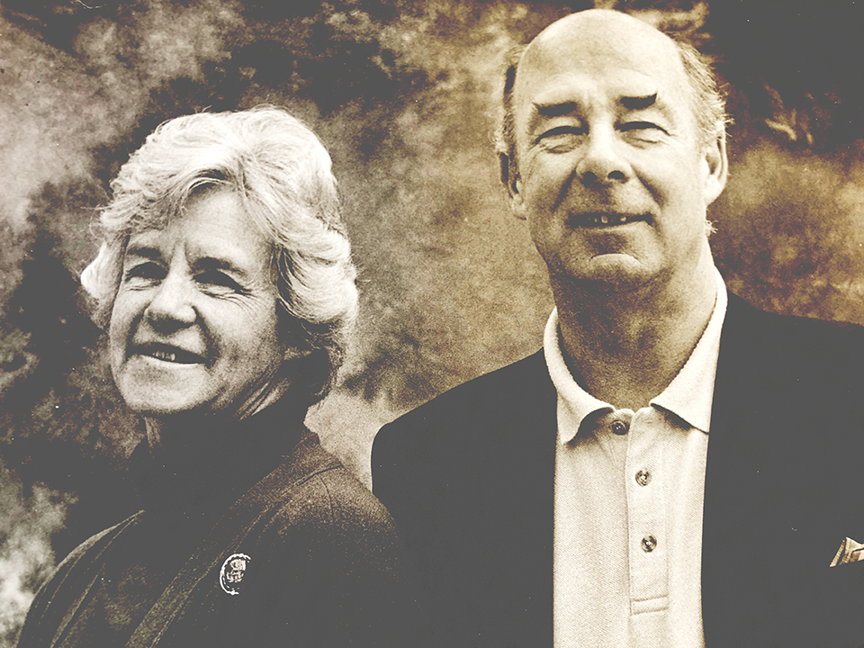Sun damaged sepia-toned portrait of an older man and woman smiling together against a dramatic cloudy background.