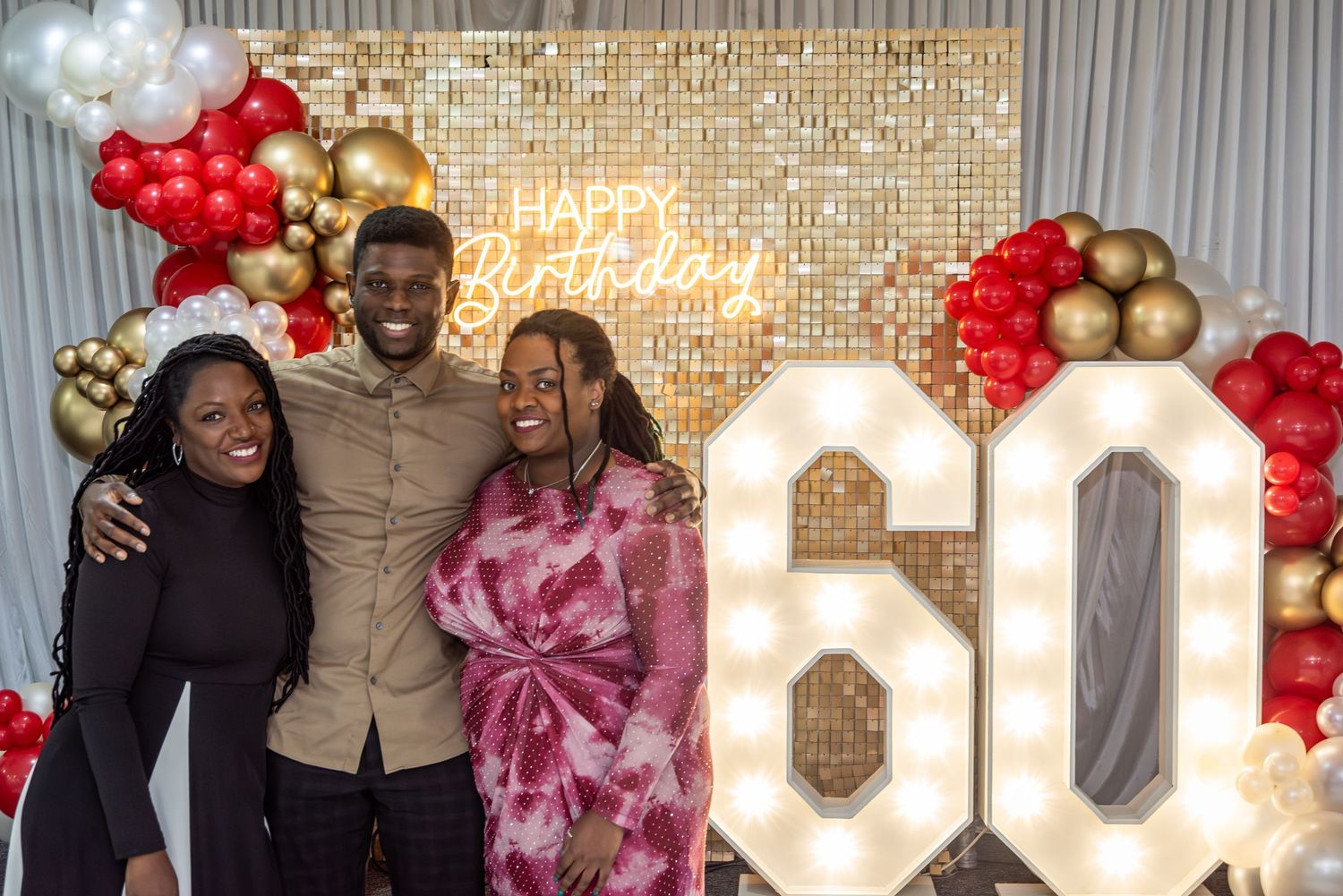 Three people posing at a 60th birthday party with balloon arch backdrop.