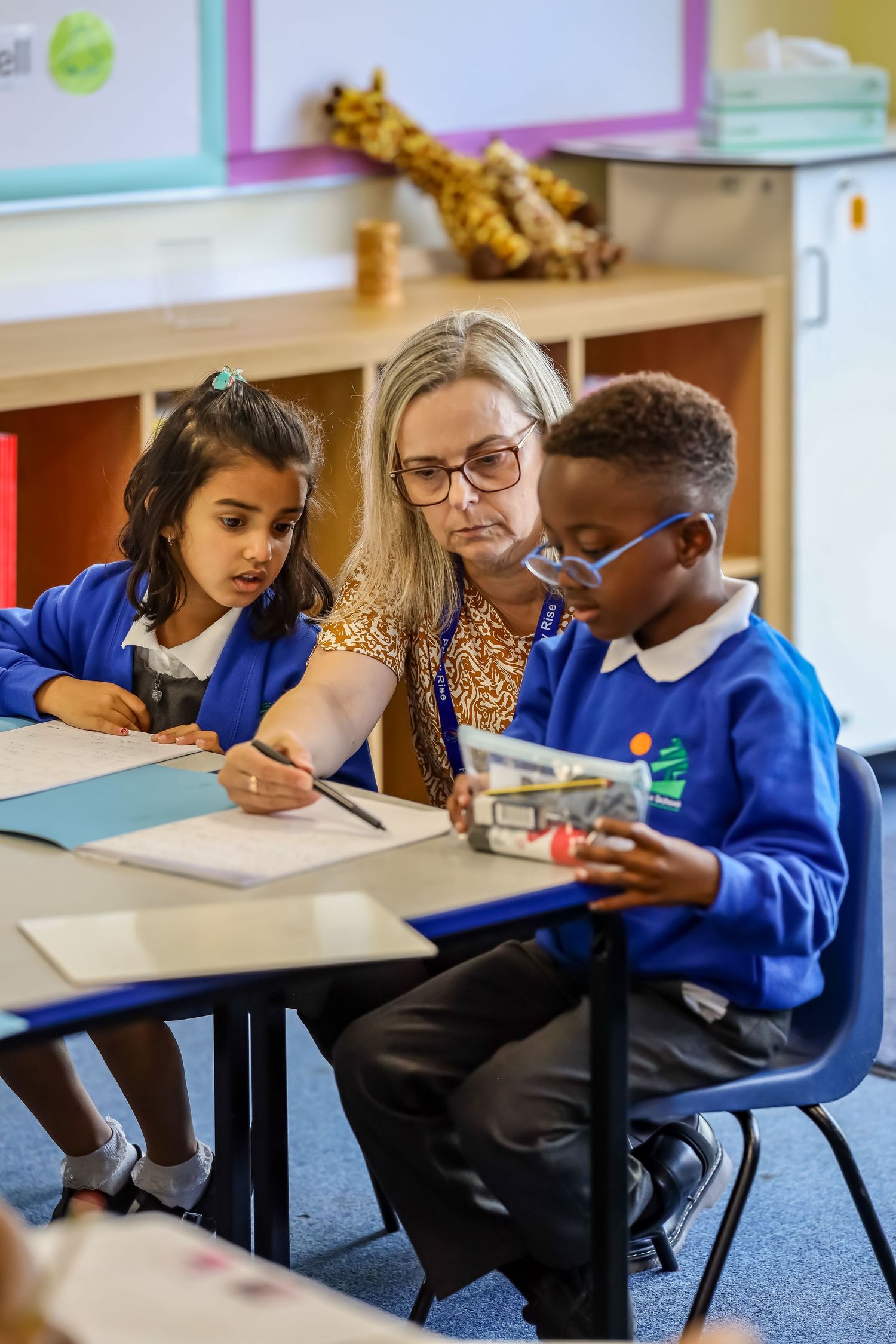 A teacher assists two young students with a writing task at a classroom desk in a primary school.